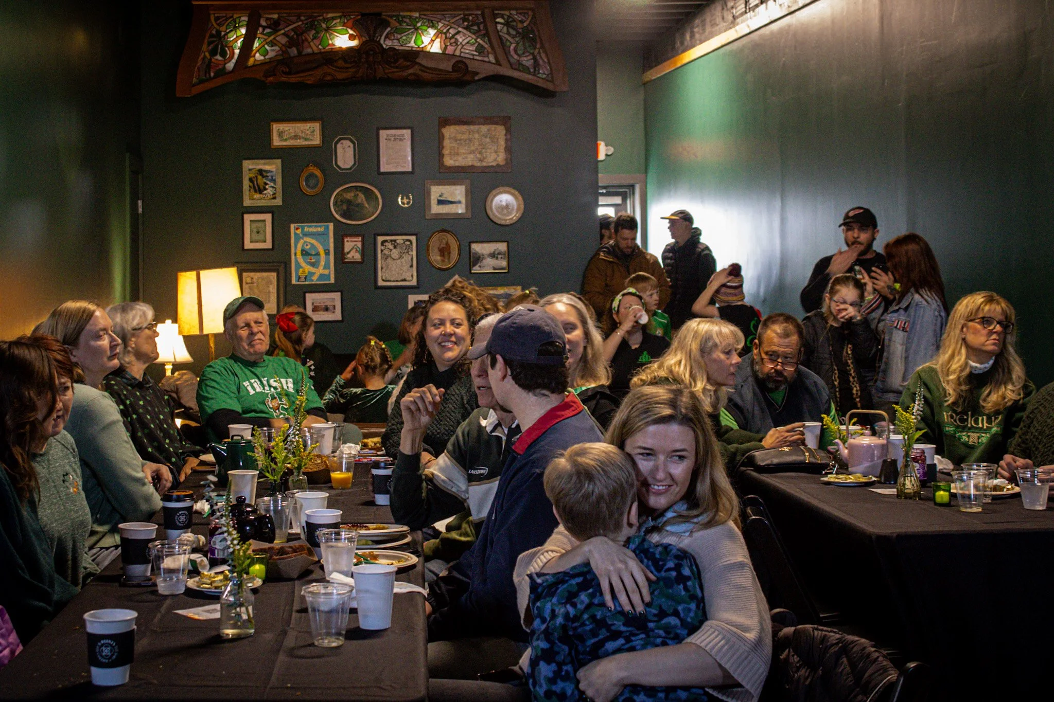 A group of people gathered in a restaurant or cafe, sitting at tables with food and drinks, with a dark green wall decorated with various picture frames and artwork behind them.