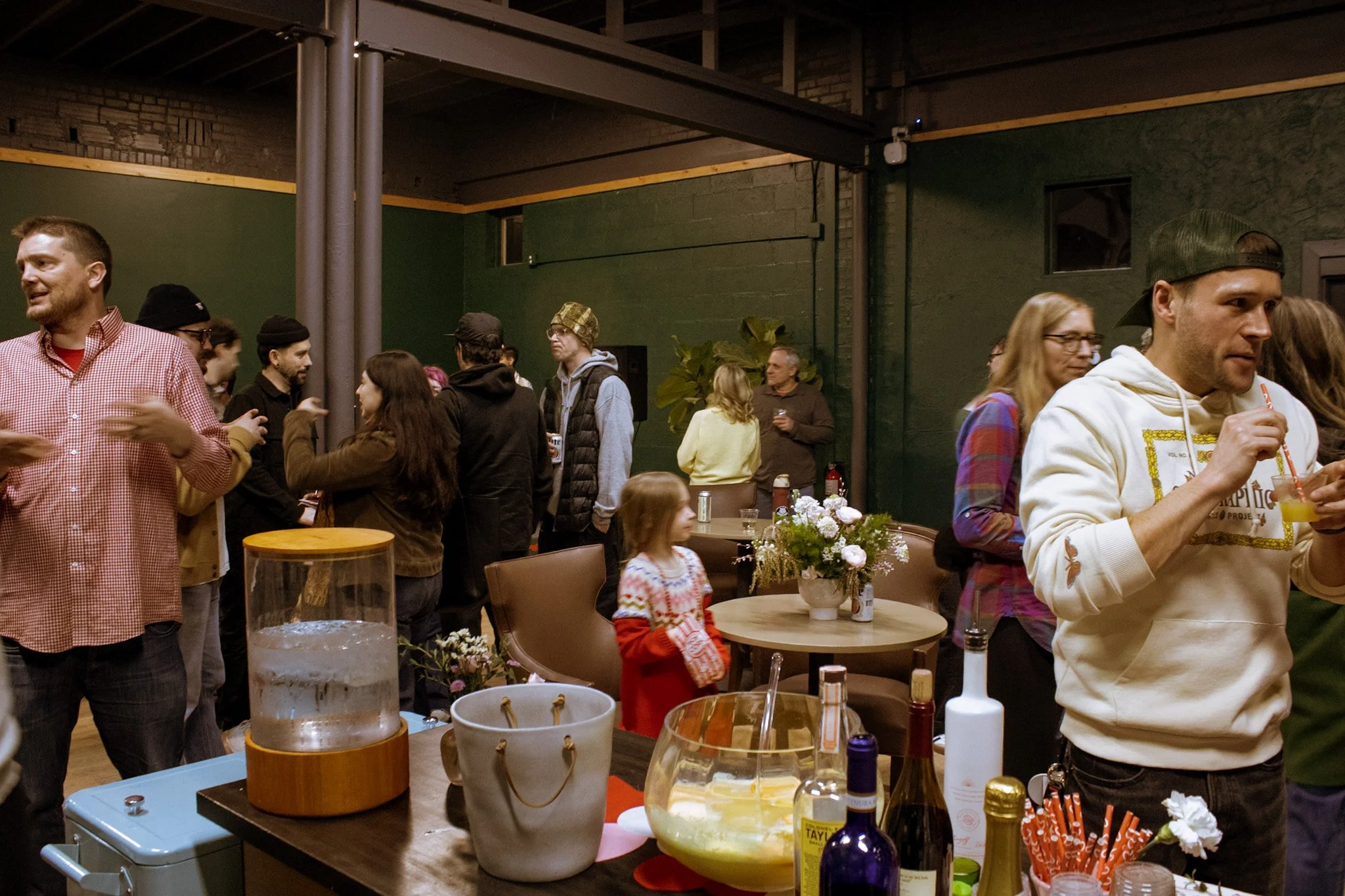 A group of people socializing at an indoor party or gathering, with various drinks and snacks on tables, and a young girl in a red dress in the foreground.