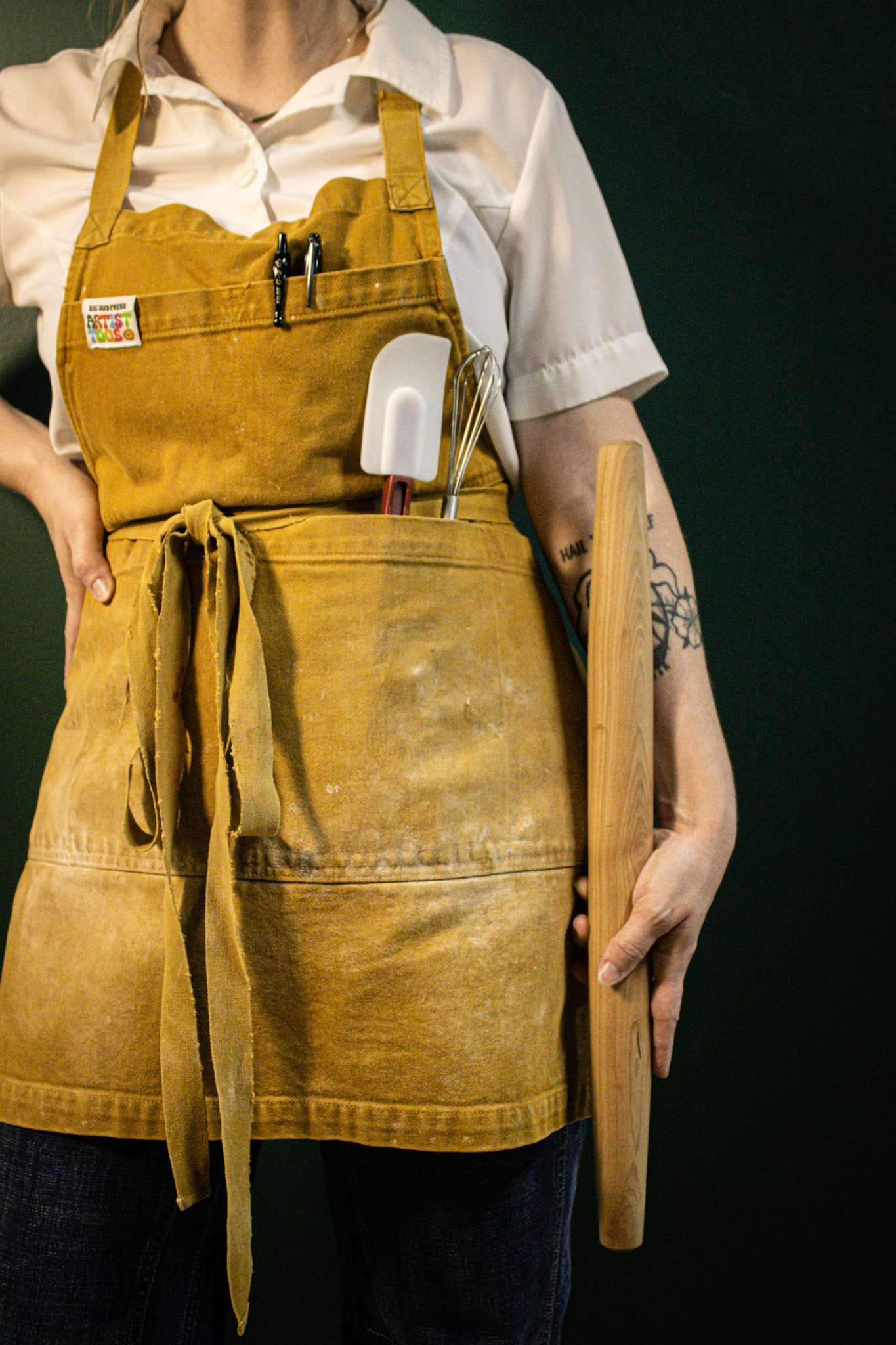 Person wearing a tan apron with tools in the pocket, holding a wooden plank, and standing against a black background.