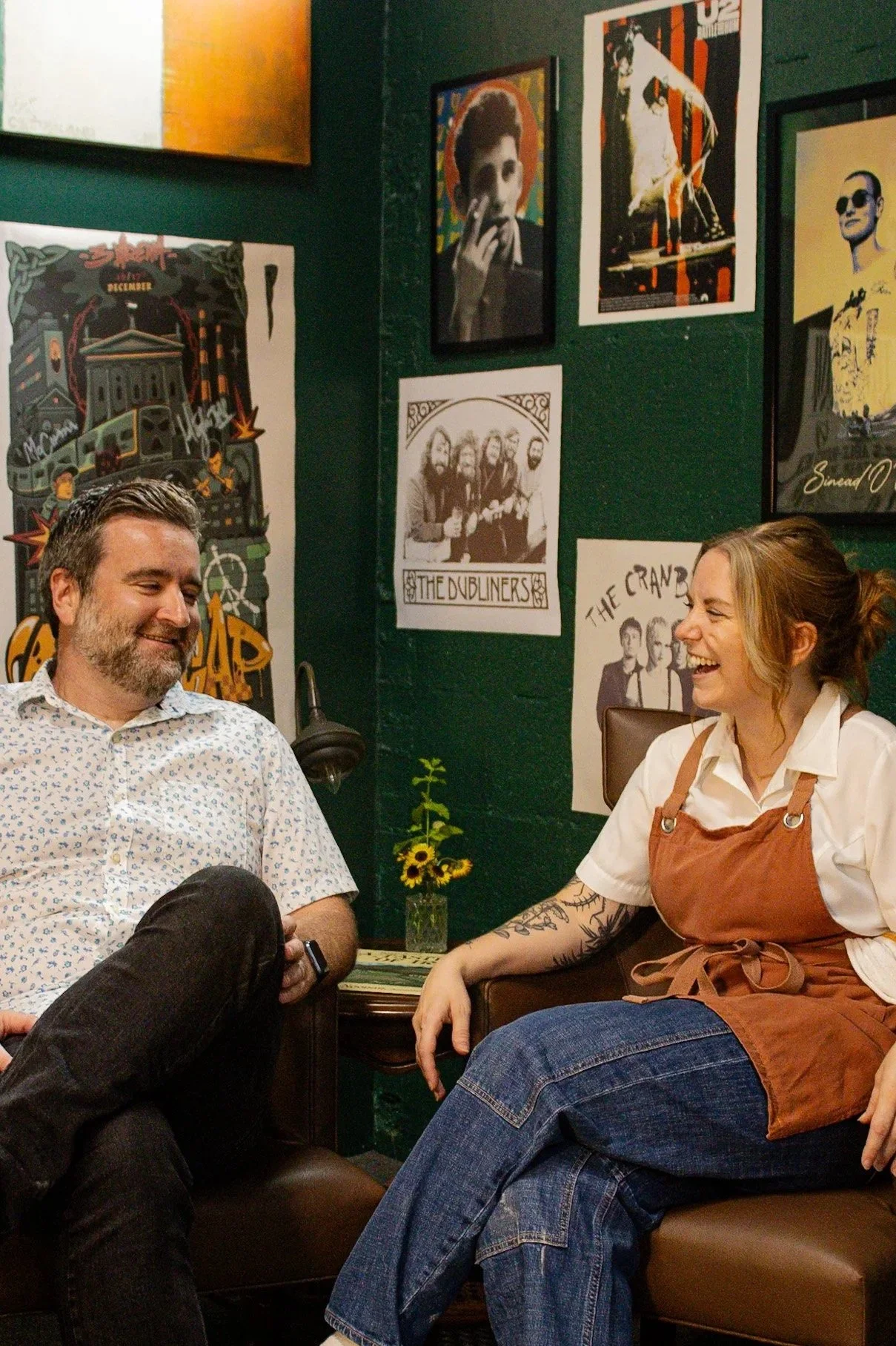 A man with gray hair and beard wearing a white shirt with a small pattern is sitting and smiling, and a woman with tattoos and red hair tied back, wearing a white shirt and brown apron, is sitting across from him, laughing. They are in a room with dark green walls decorated with music and pop art posters, including images of musicians and bands.