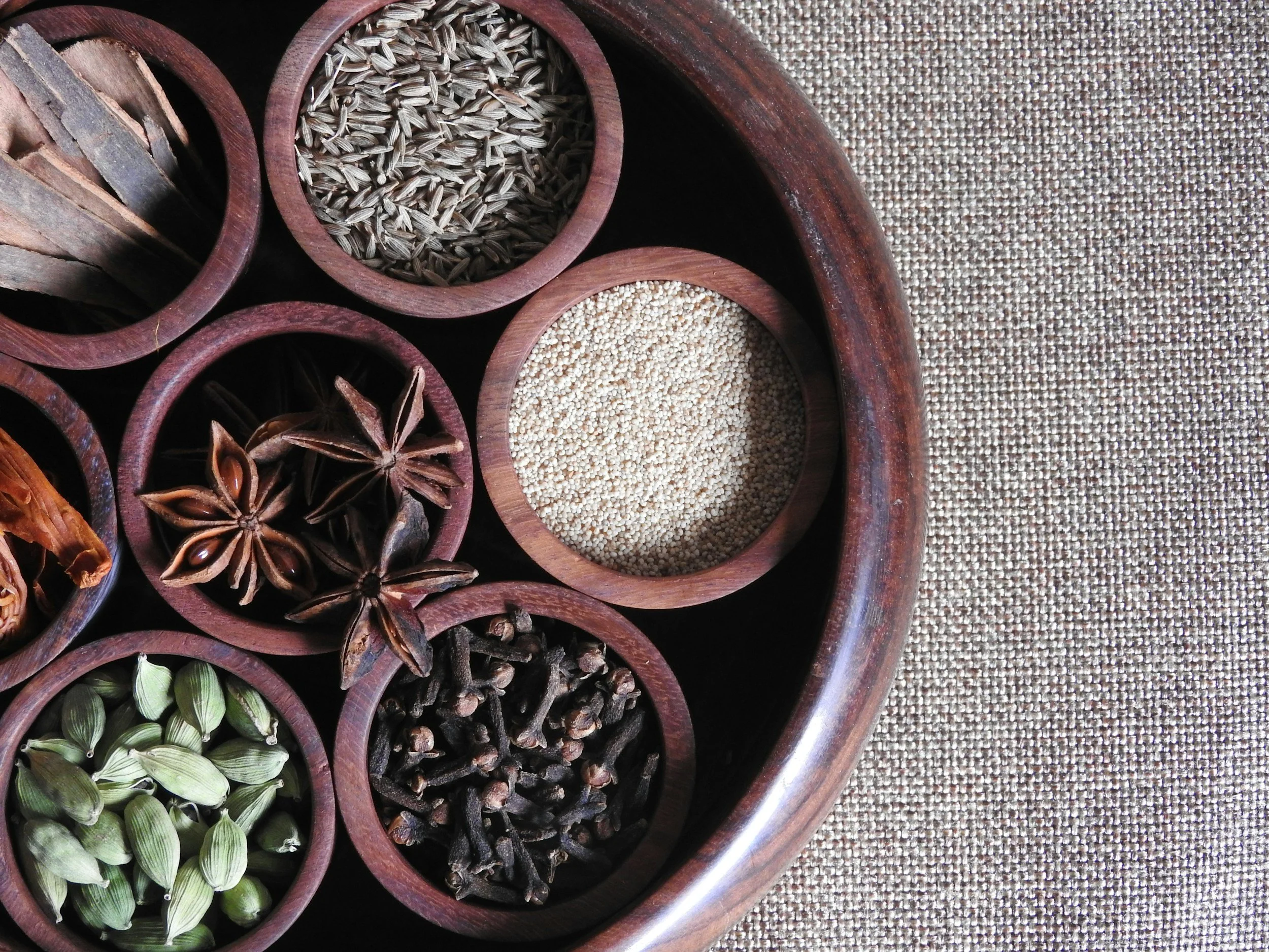 Top view of a round wooden container with multiple small compartments holding various spices and seeds, placed on a textured surface.