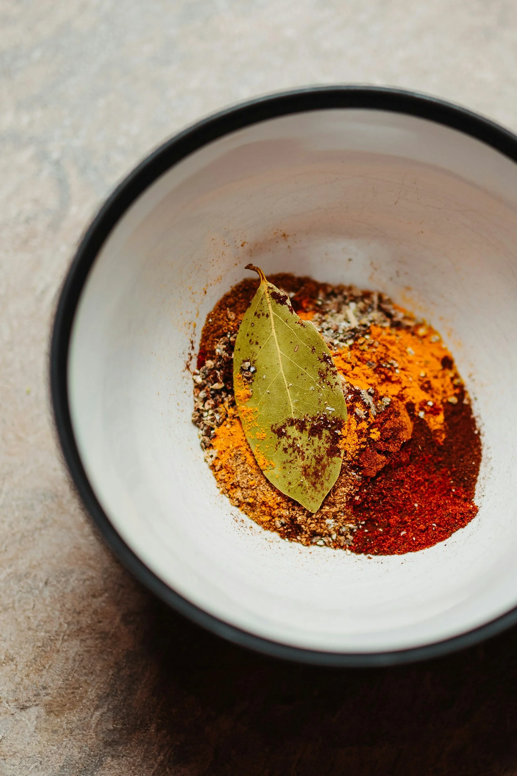 A top view of a mug containing herbs and spices, including bay leaf, turmeric powder, black pepper, and chili powder, on a textured gray surface.