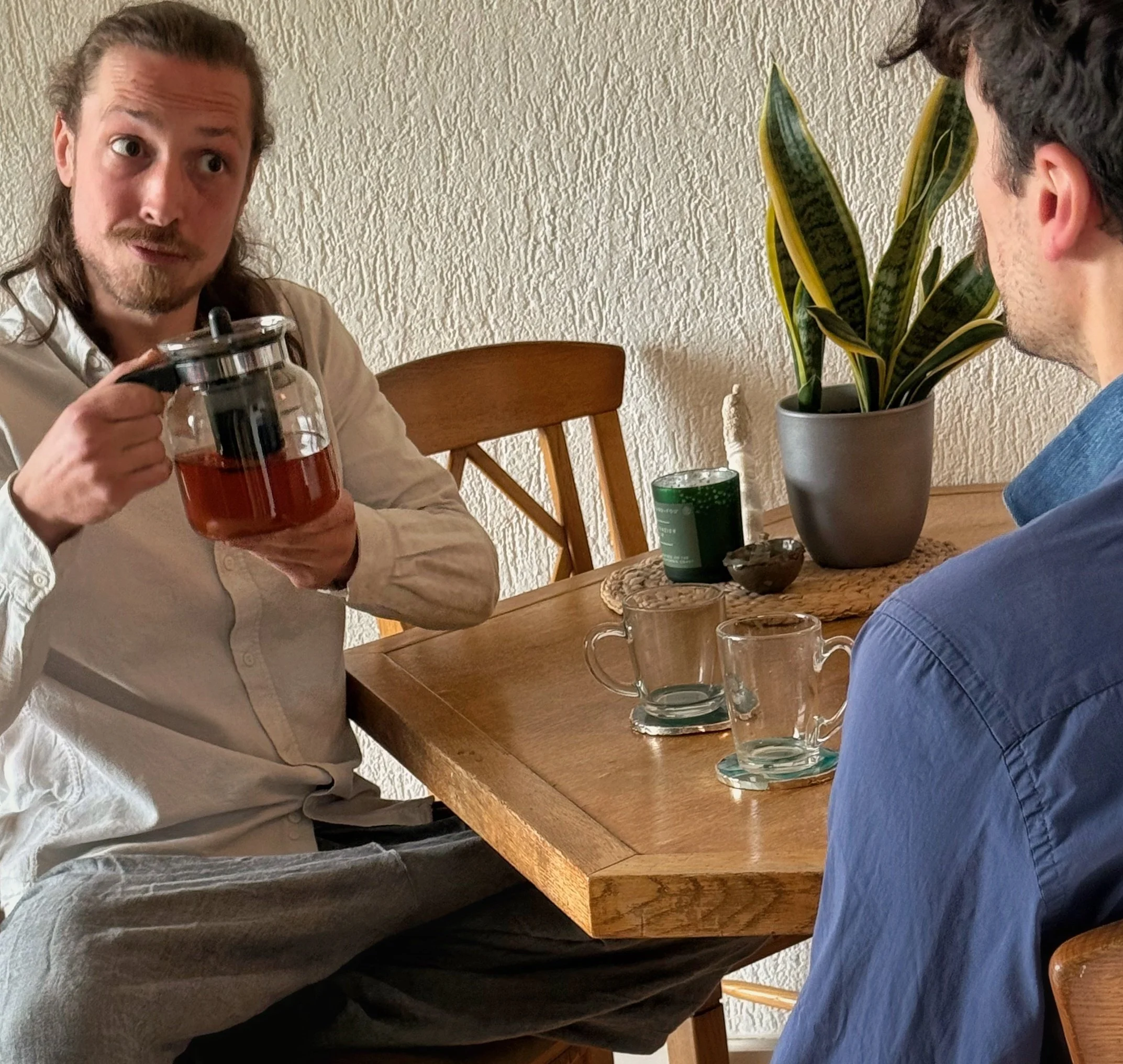 Twee mannen zitten aan een houten eettafel, één is scherp in beeld en houdt een thee kan vast. De ander zit met zijn rug naar de camera. Op de tafel staan verschillende glazen en een groen blikje. Achter hen staat een grote plant in een pot.