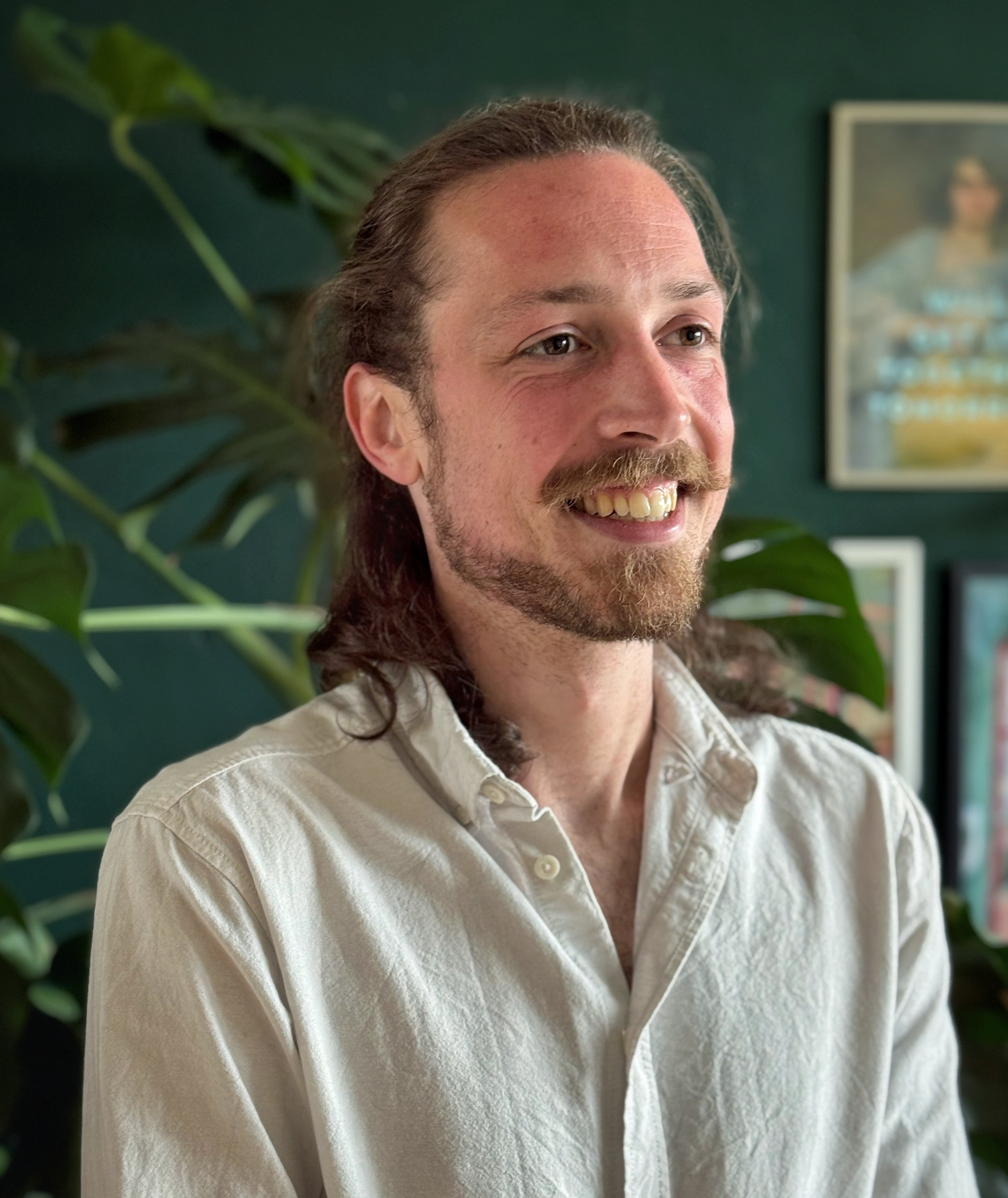 Portret van een lachende man met lang, bruin haar en een lichte baard, gekleed in een lichte, crèmekleurige overhemd, staand voor een donkergroene muur met een foto en omringd door groene bladeren.