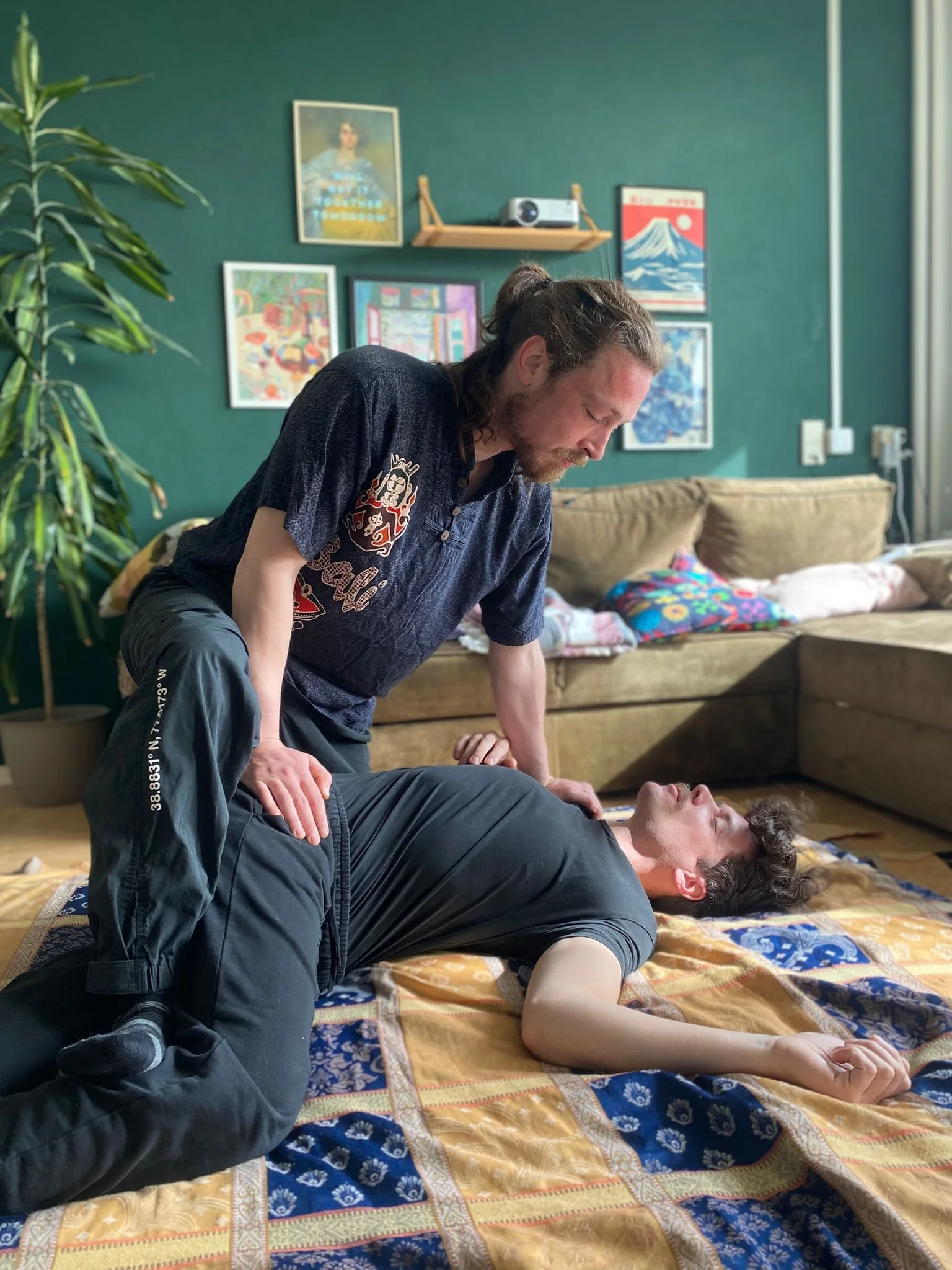 Een man ligt op zijn rug op een mat in een woonkamer terwijl een andere man boven hem knielt en zijn hand op zijn schouder legt voor een strekking tijdens een shiatsu behandeling.