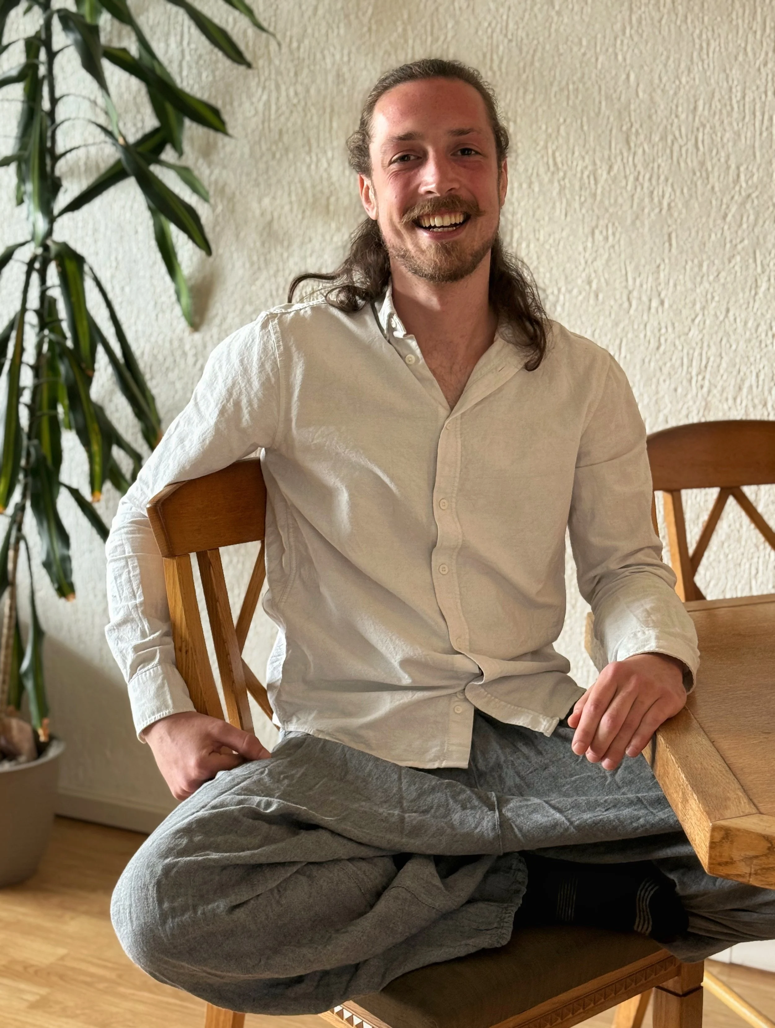 Man met lang haar en baard lachend zittend op een stoel, aan tafel, in een huis met houten vloer, naast een groene plant.