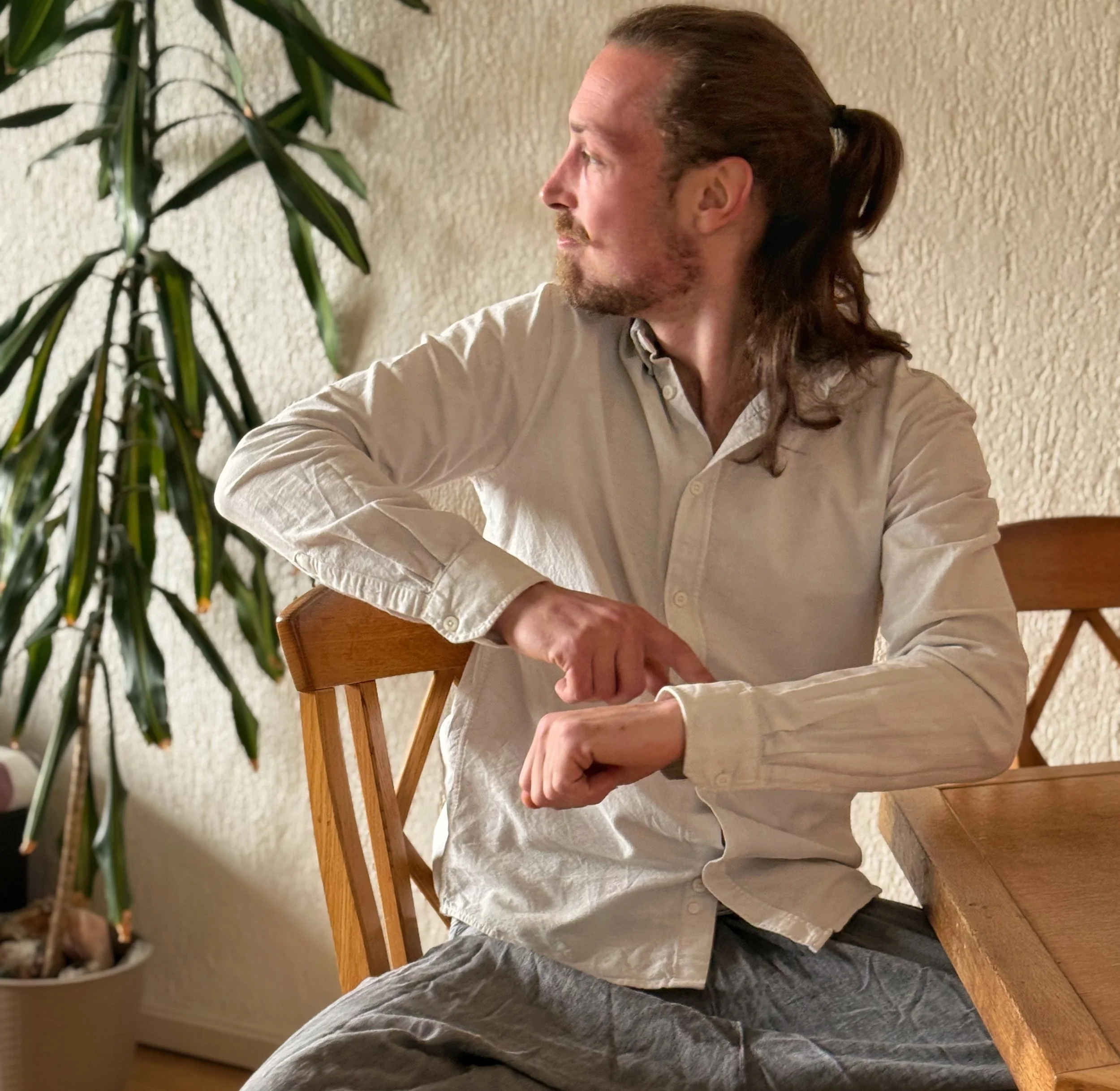 Man met lang haar in paardenstaart zit aan een houten tafel, met een groene plant op de achtergrond.
