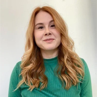 A young woman with long, wavy red hair wearing a green top, standing against a plain white background.