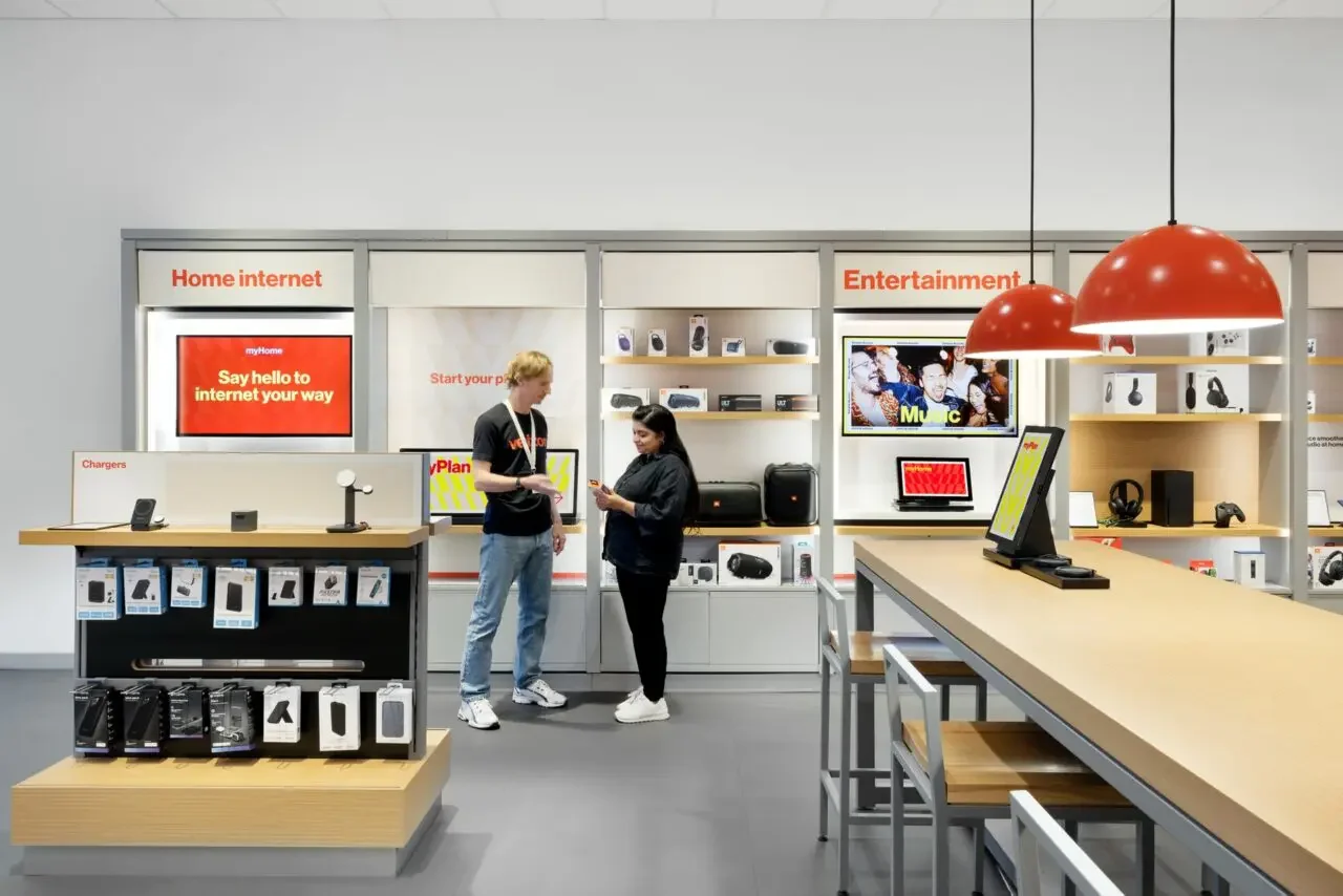 Two people shopping for electronics in a store with sections labeled 'Home internet' and 'Entertainment', browsing digital devices and accessories on display.