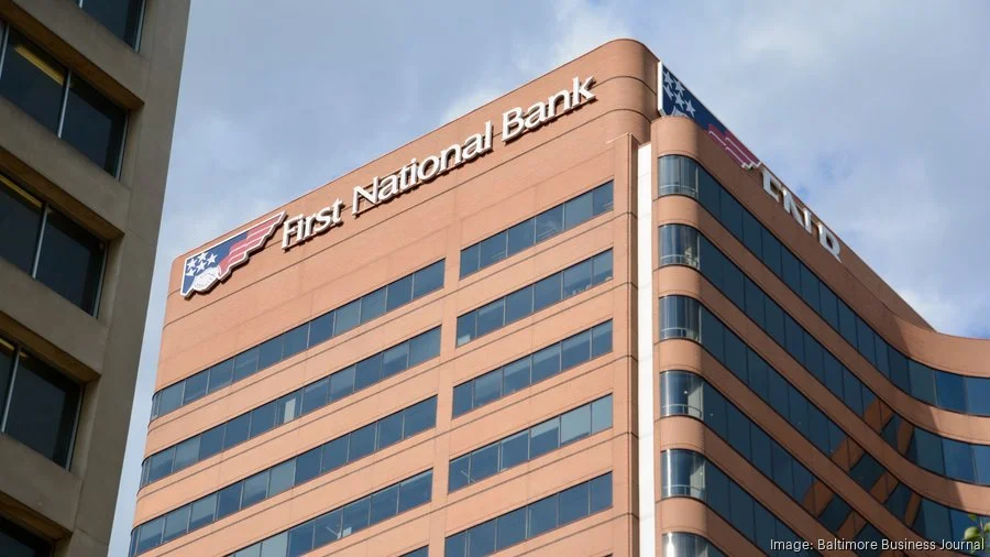 A tall modern office building with the sign 'First National Bank' at the top corner. The building has horizontal rows of windows and rounded edges on one side, with a blue and red American flag emblem near the sign.