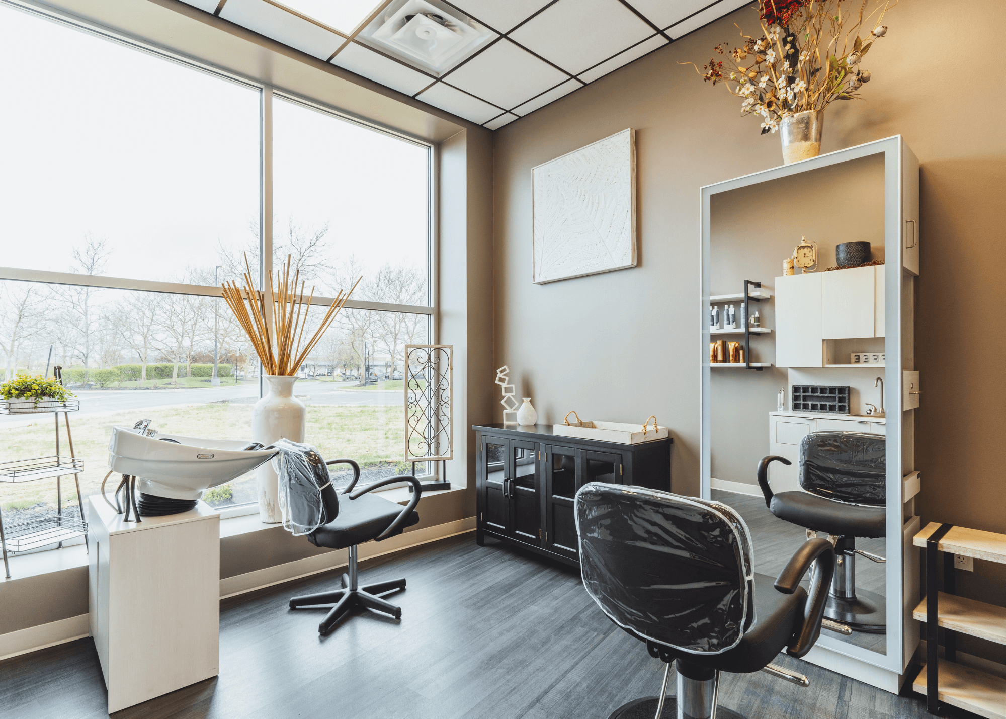 A modern hair salon interior with large windows, a mixing sink, black salon chairs, a white wall cabinet with decorative items, and a large vase with dried flowers on top.