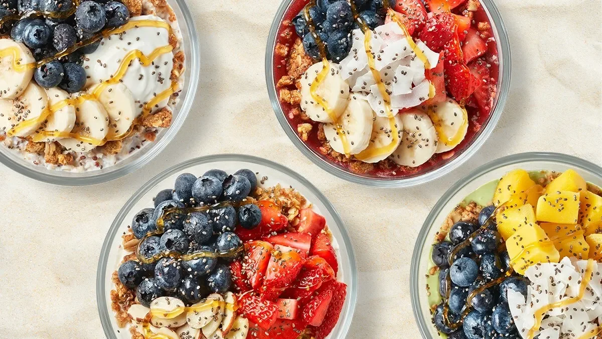 Four bowls of fruit parfaits topped with blueberries, strawberries, bananas, mango, coconut flakes, chia seeds, and honey.