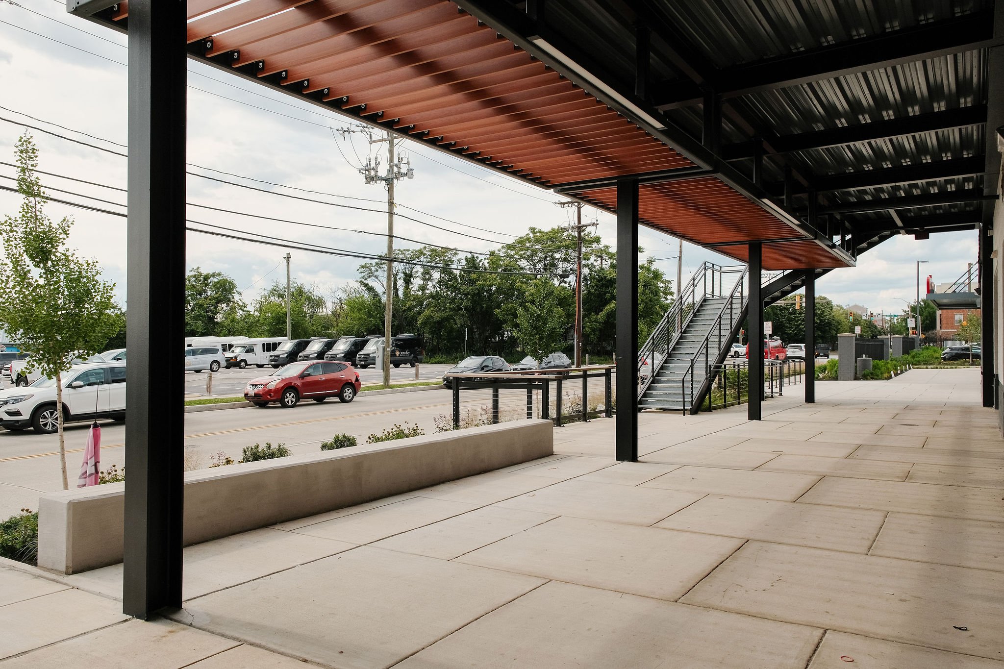 View from under a building with a modern architectural design, featuring a black and wooden ceiling structure, stairs leading up, and a parking lot with cars and trees in the background.