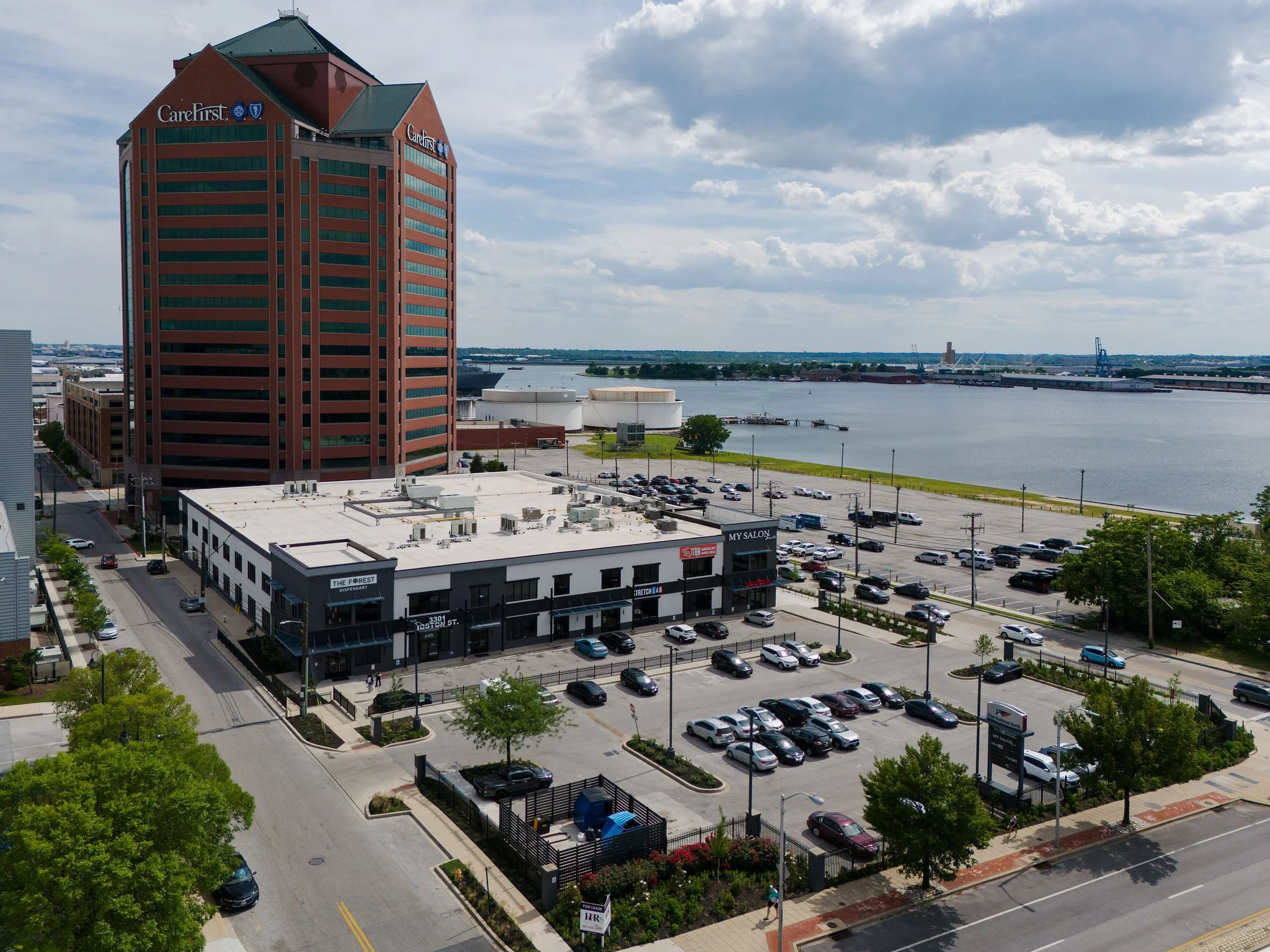 An aerial view of a cityscape near a body of water, featuring a tall red-brick office building with green windows, a parking lot with many cars, and several smaller commercial buildings. The scene includes a cloudy sky and a distant shoreline with industrial cranes.