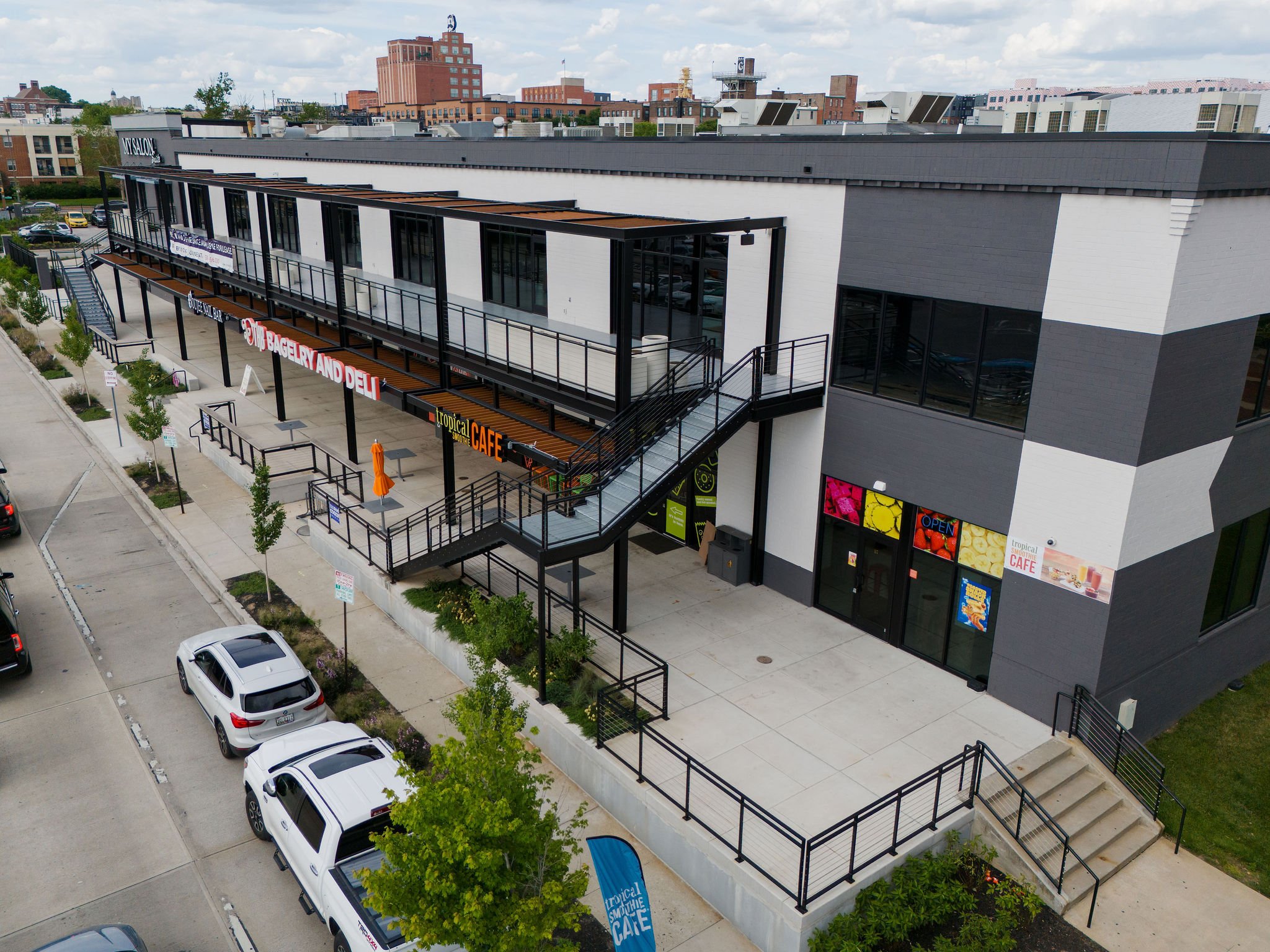 A modern, multi-story commercial building with a black metal railing on a balcony and stairs outside, parking lot with several cars, small trees, and colorful banners advertising a café.
