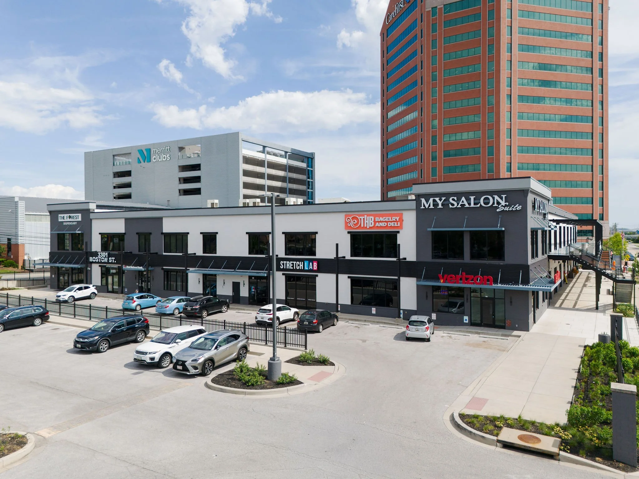 A parking lot with several parked cars in front of a commercial building and tall office buildings in the background under a partly cloudy sky.