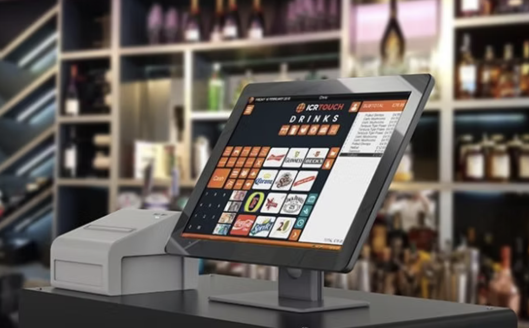 A modern touchscreen point-of-sale register displaying a drinks menu in a bar or restaurant, with a cash drawer underneath, against a blurred background of shelves with liquor bottles and bar supplies.