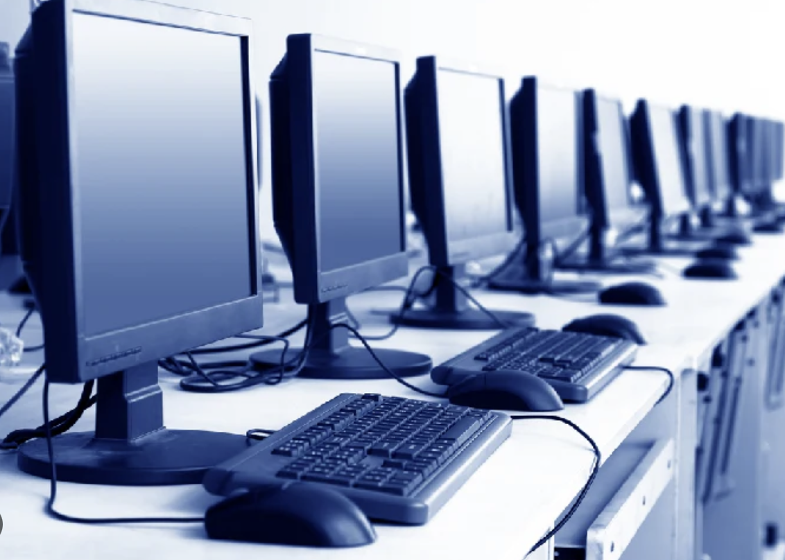 Row of computer workstations with monitors, keyboards, and mice on a long table in a computer lab or office setting.