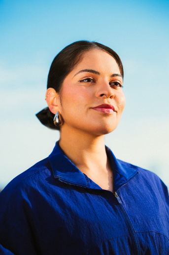 A woman with dark hair tied back, wearing a blue jacket and earrings, against a cloudy sky background.