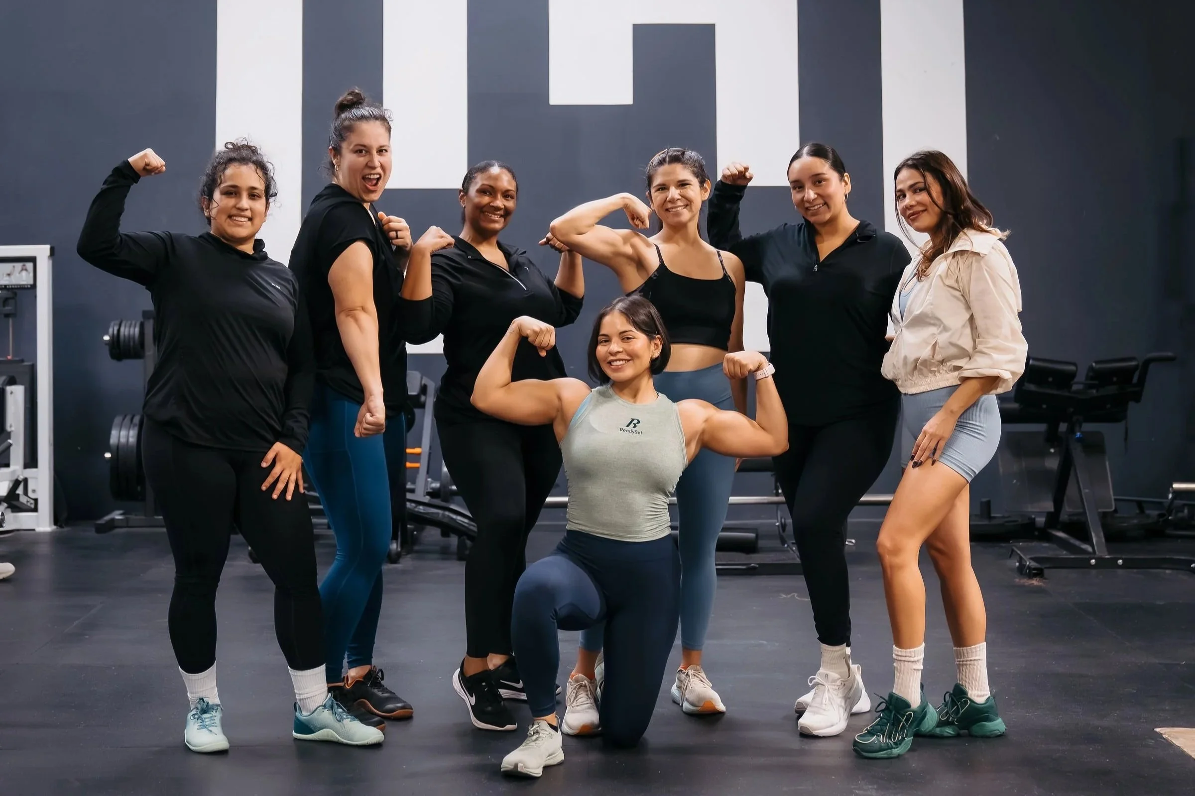 Group of seven women in gym clothes posing with flexed arms and smiling, showing strength and confidence, inside a gym with weightlifting equipment and a dark wall background.