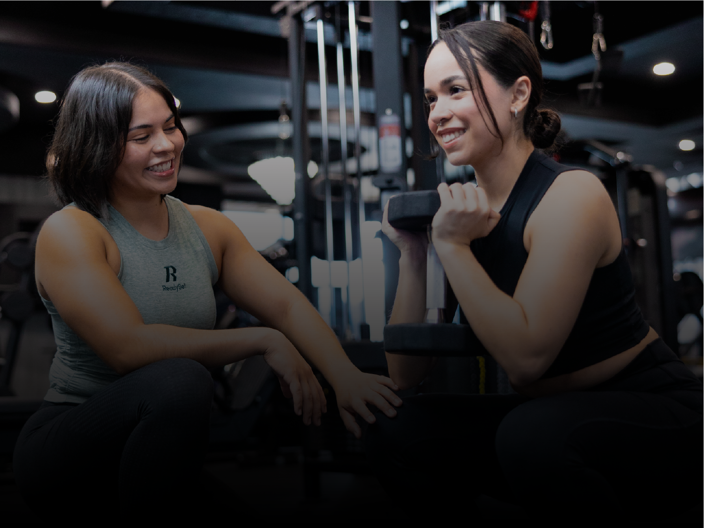 Two women working out in a gym, one spotter is assisting the other with a dumbbell exercise, smiling and encouraging.