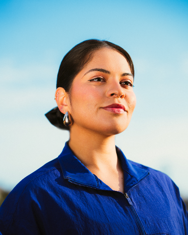 A young woman with dark hair pulled back, wearing a blue jacket and earrings, outdoors against a blue sky.