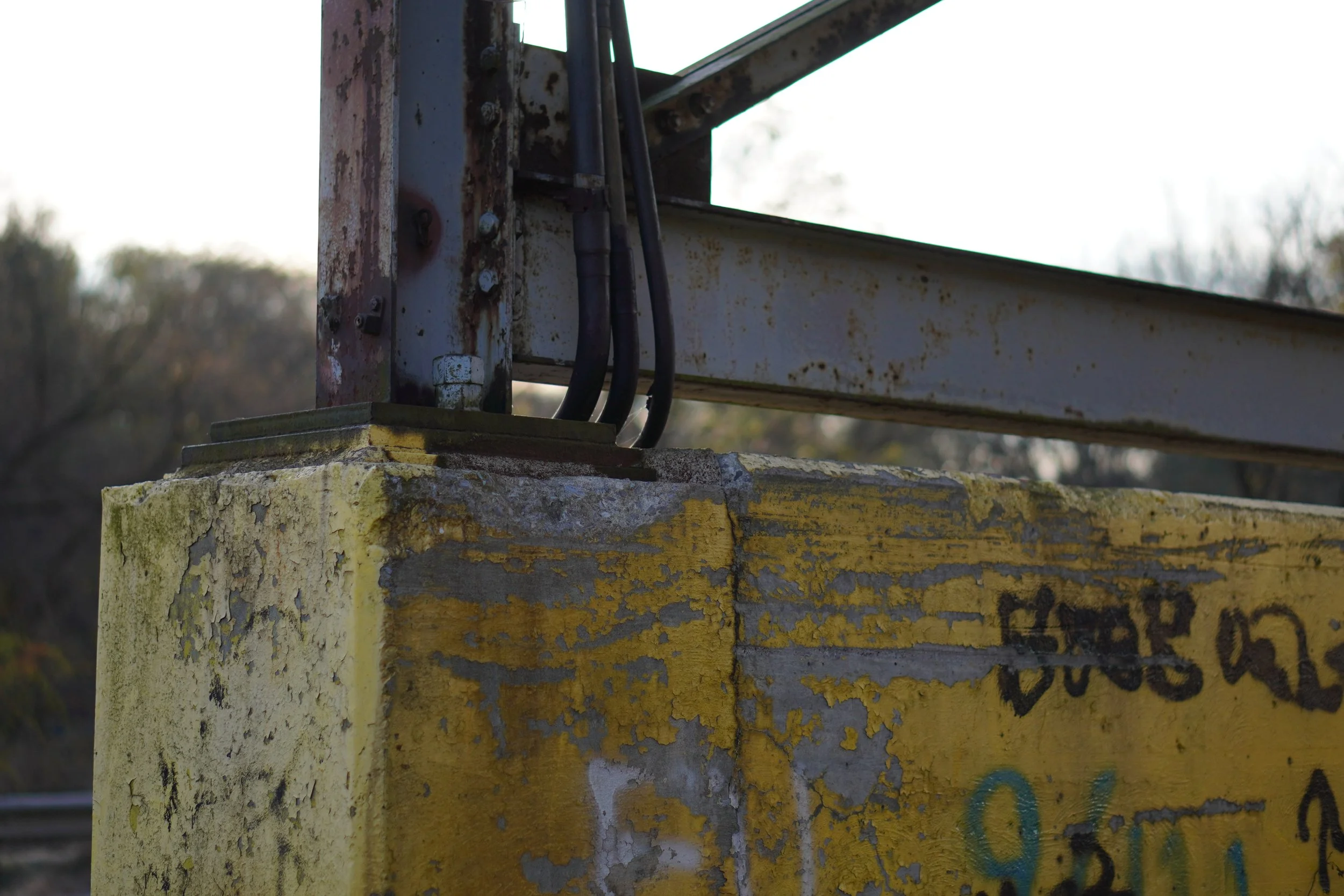 Close-up of a weathered, rusty metal structure mounted on a concrete base with peeling yellow paint and graffiti, with a blurry background of trees and sky.