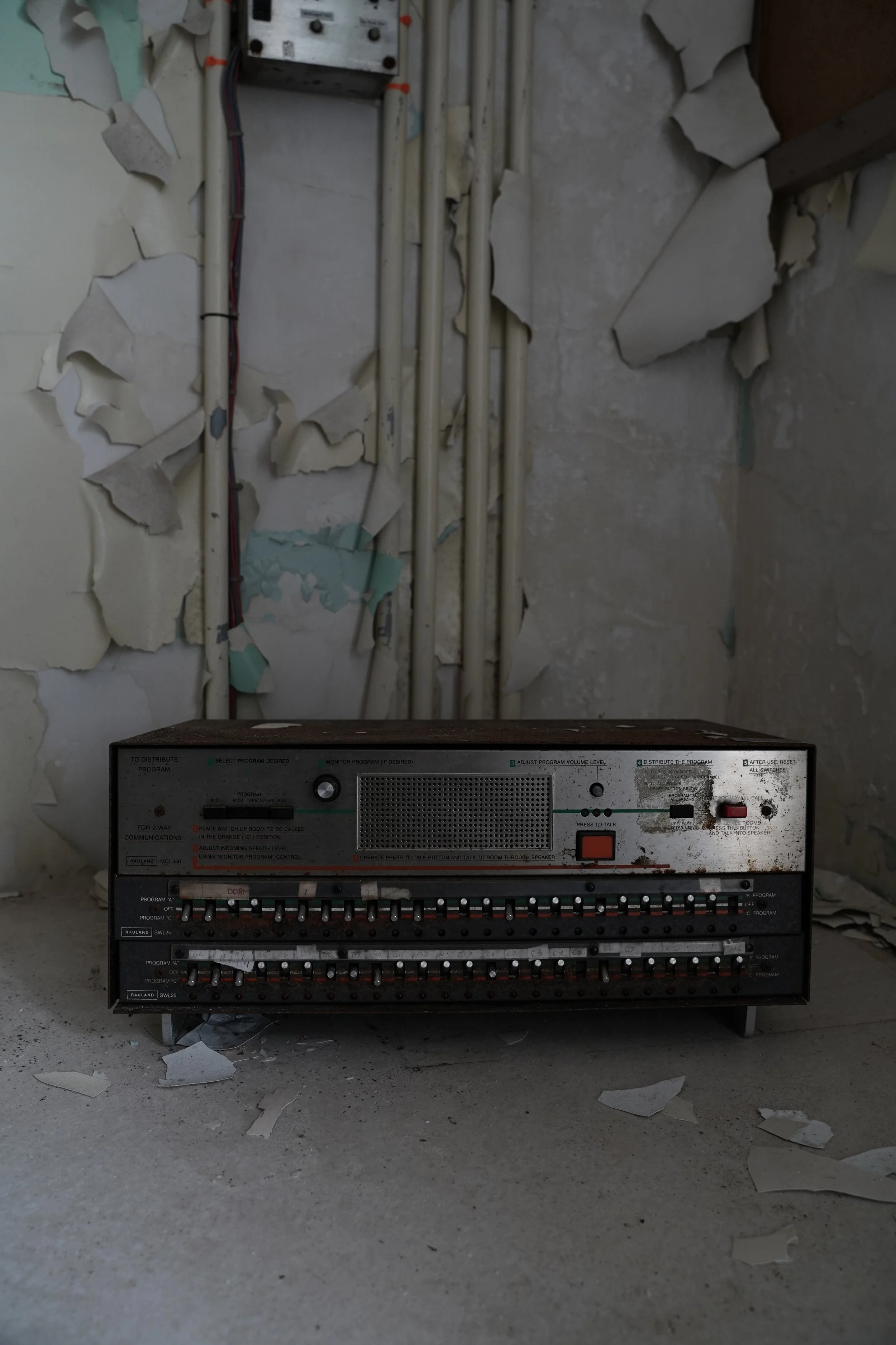 Old electronic control panel with numerous switches and buttons, sitting on a dusty floor in front of a peeling wall with exposed pipes.