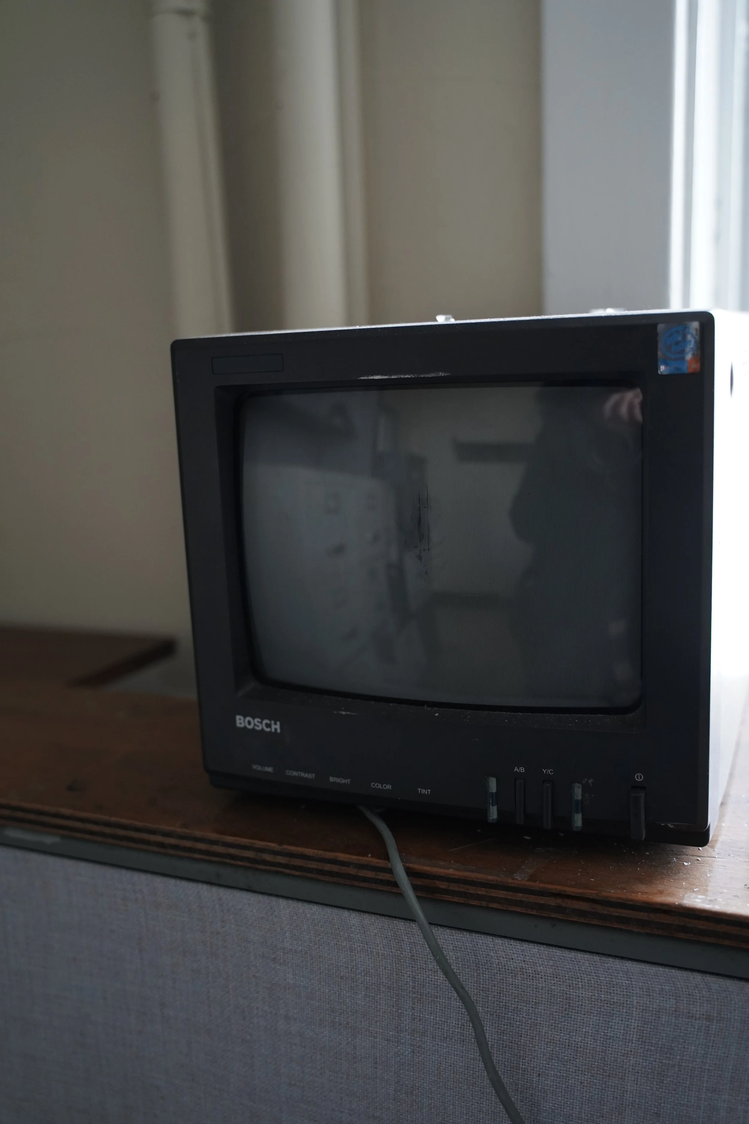 A vintage black Bosch monitor displaying a blank screen, placed on a wooden desk, with a cord hanging down in front of it. The background features a plain wall and a window.