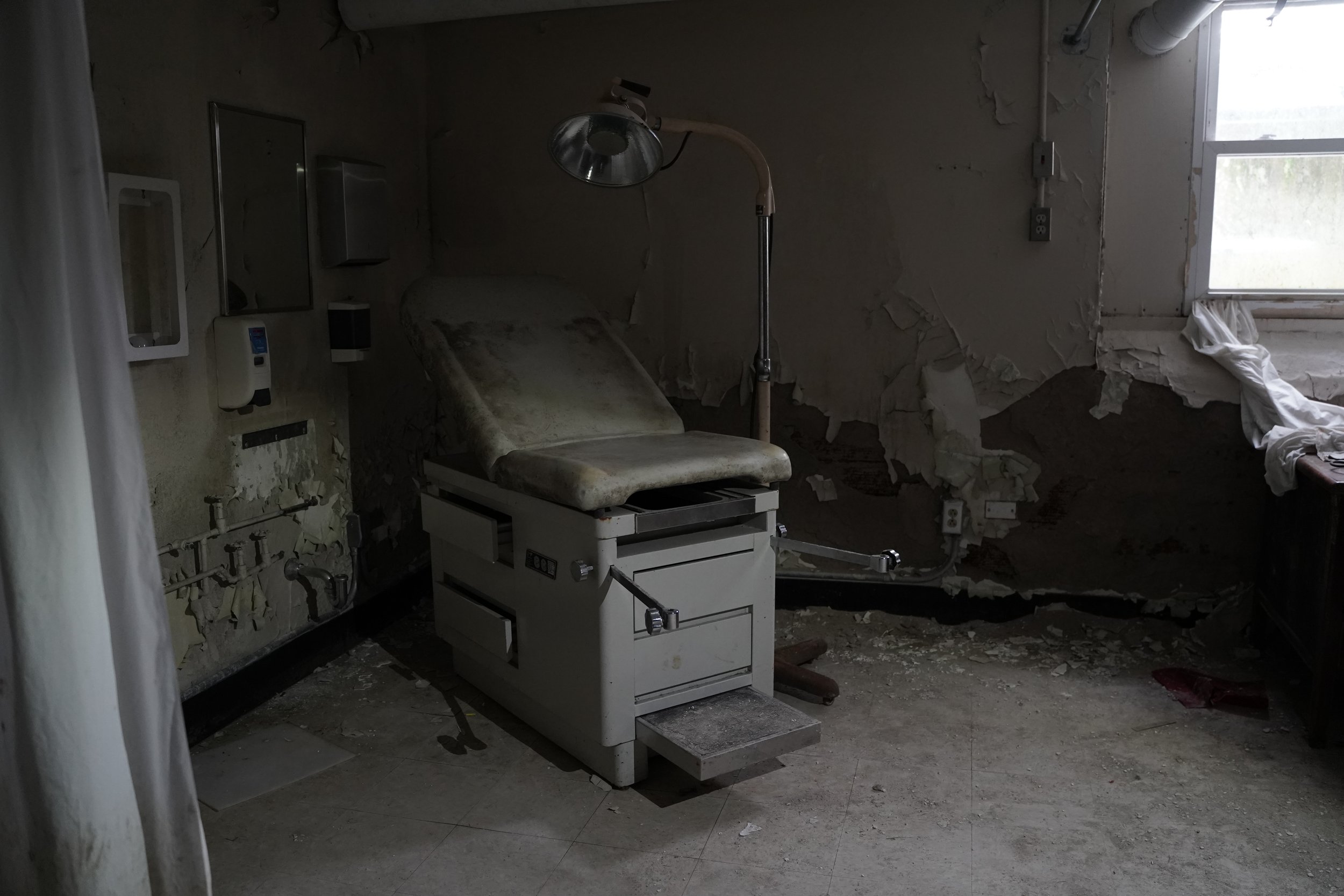 A dilapidated medical room with peeling walls, a worn examination table, and medical equipment scattered around.
