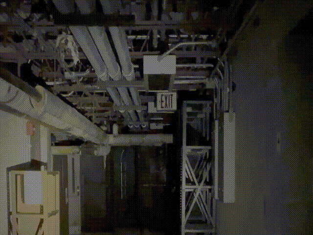 A dark industrial hallway with exposed pipes and metal shelving units, an illuminated exit sign hanging from the ceiling.