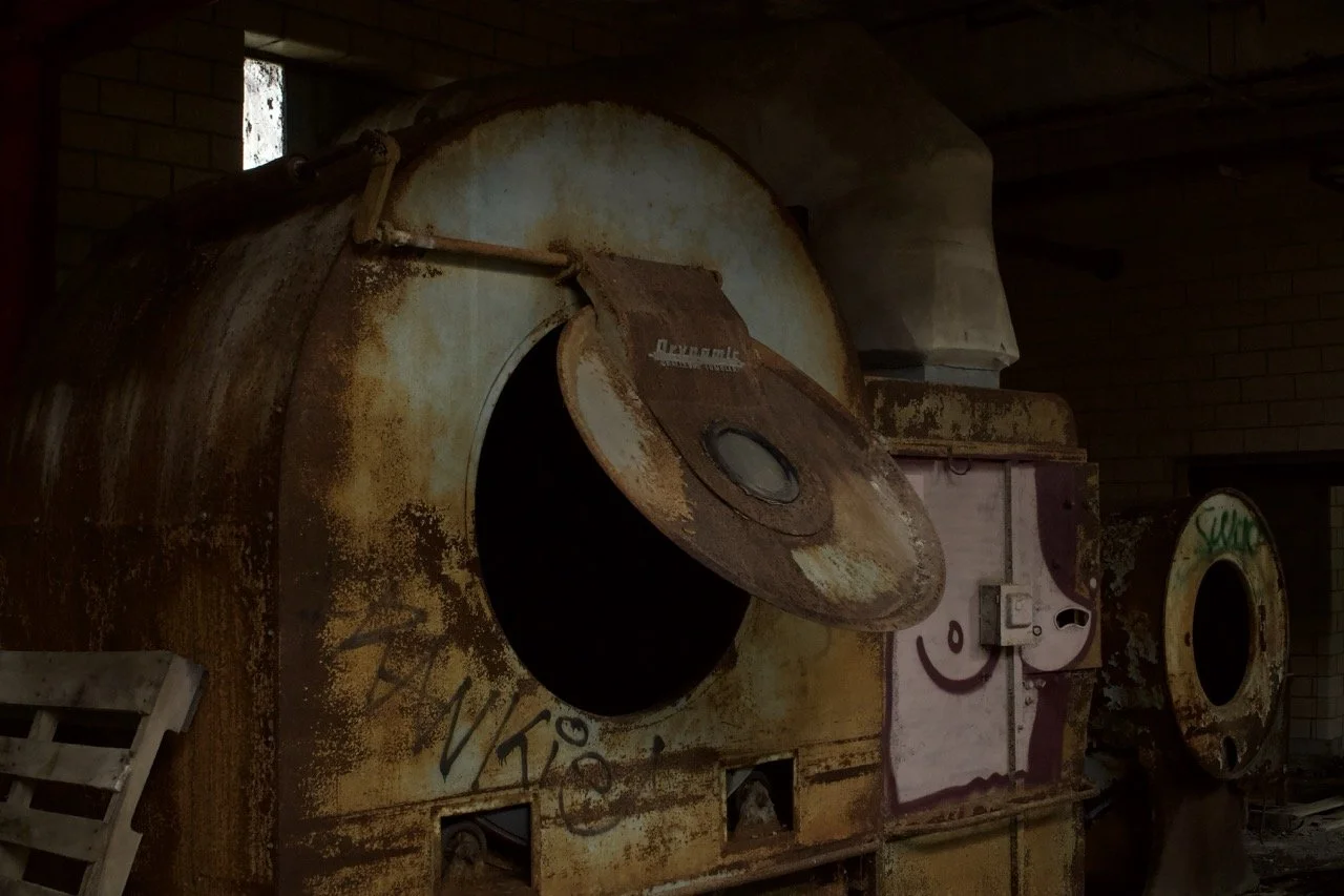 An abandoned, rusty industrial washing machine with its door open, located in a dimly lit, possibly abandoned building.