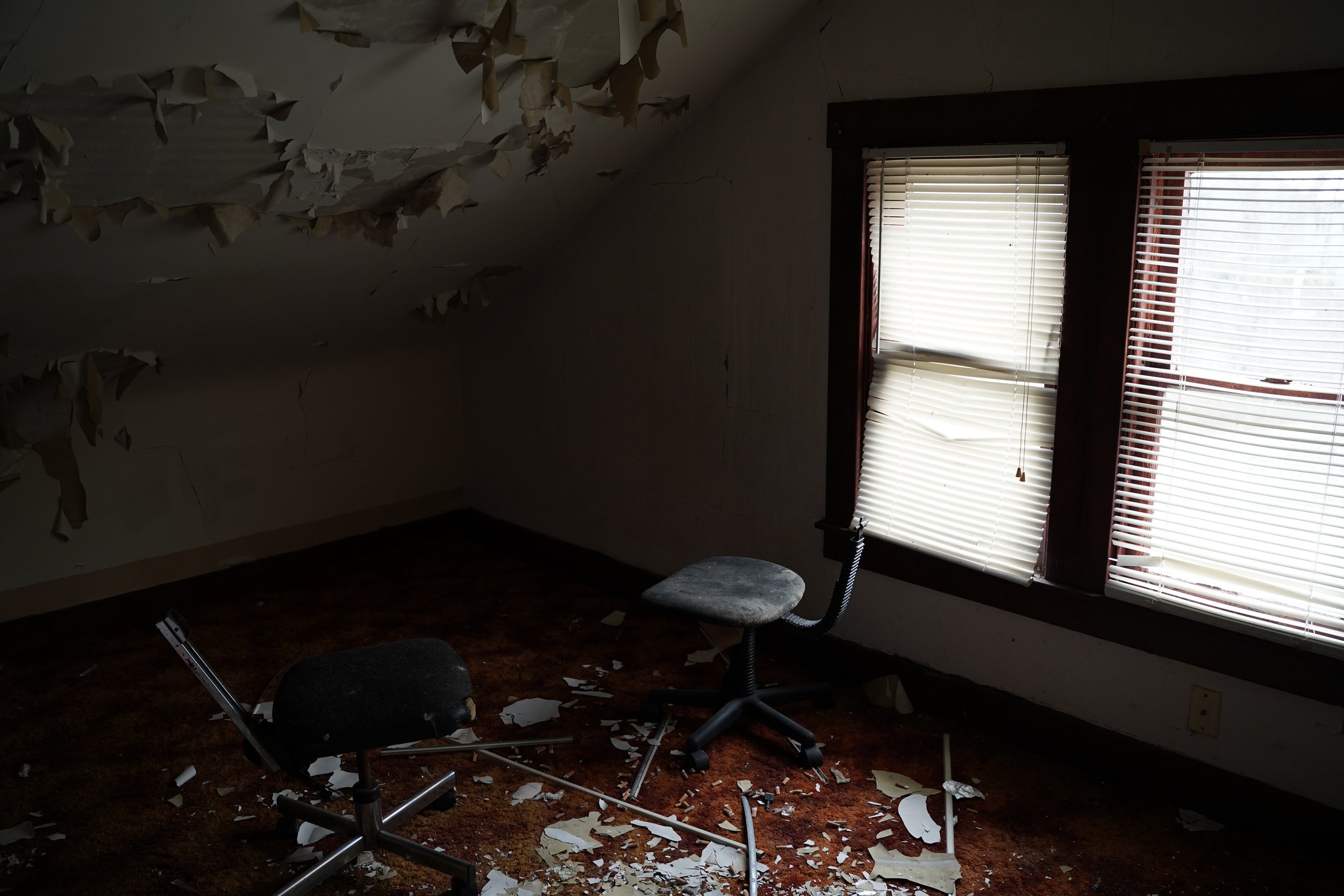 A room with a damaged ceiling and broken window blinds. Two office chairs, one upright and one tipped over, sit on a carpeted floor covered with fallen ceiling material and debris.