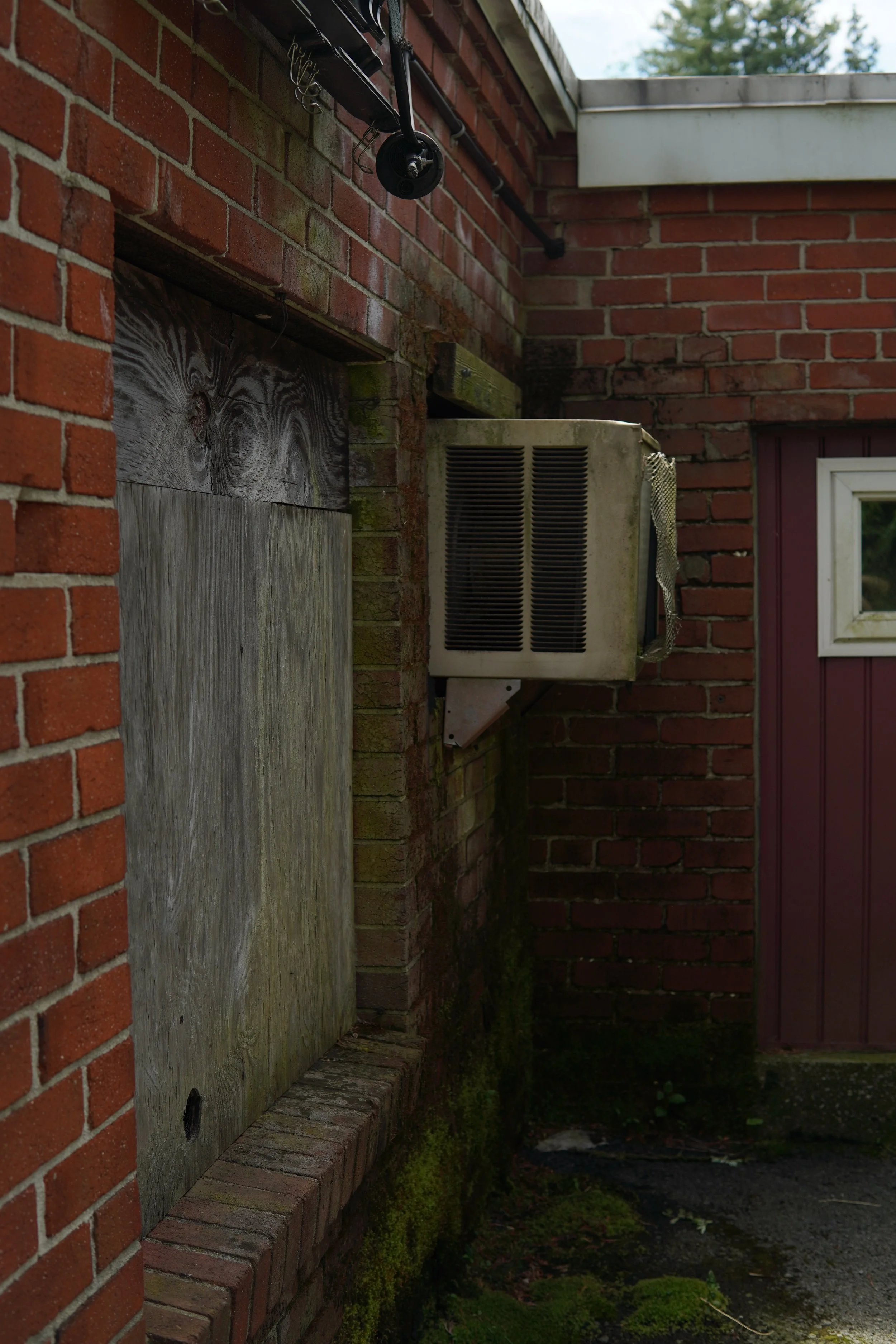 Outside brick wall with weathered wooden panel and moss, air conditioning unit, and red door with window, on a cloudy day