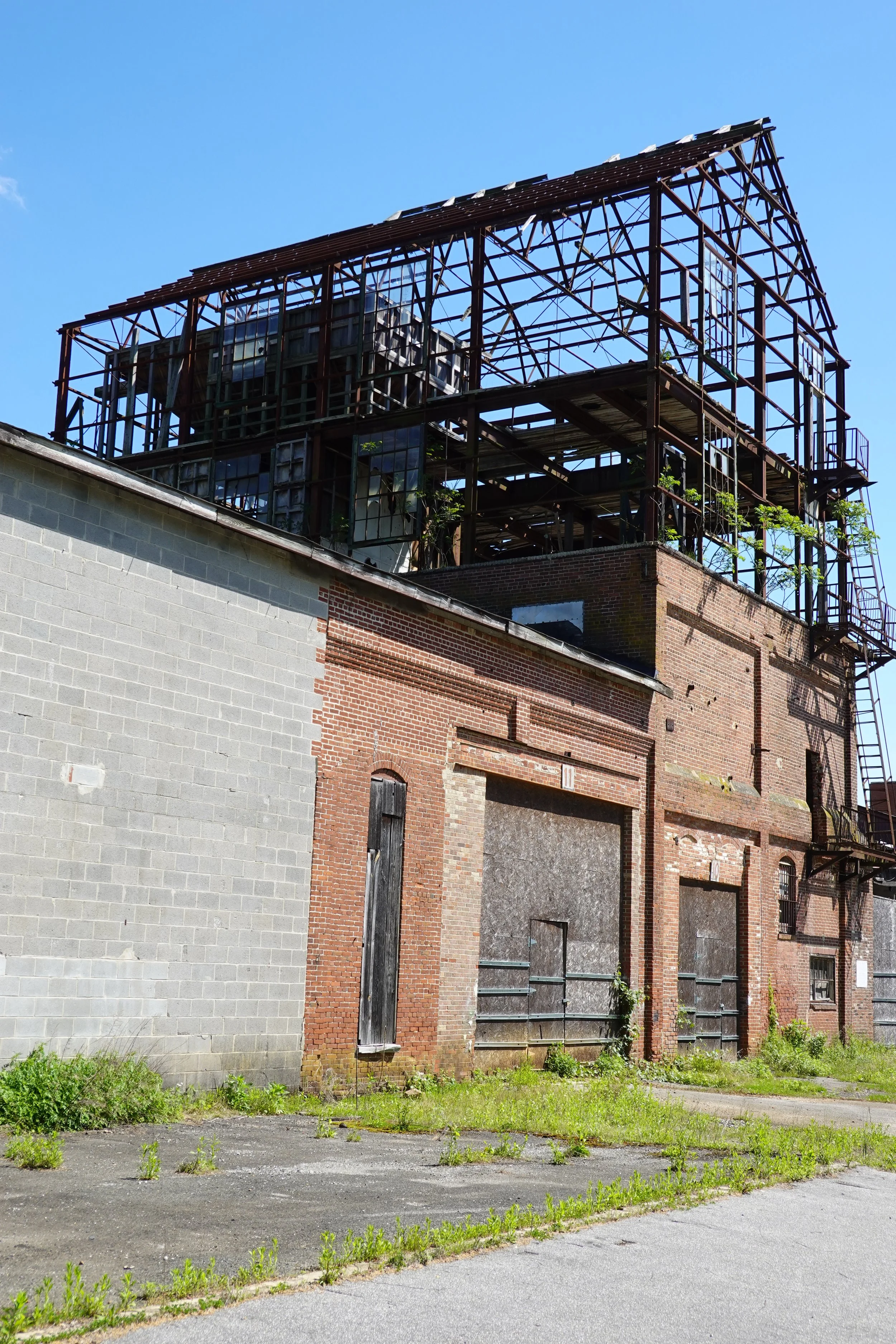 An abandoned brick building with boarded-up windows and a metal framework on the roof, surrounded by overgrown grass and weeds.