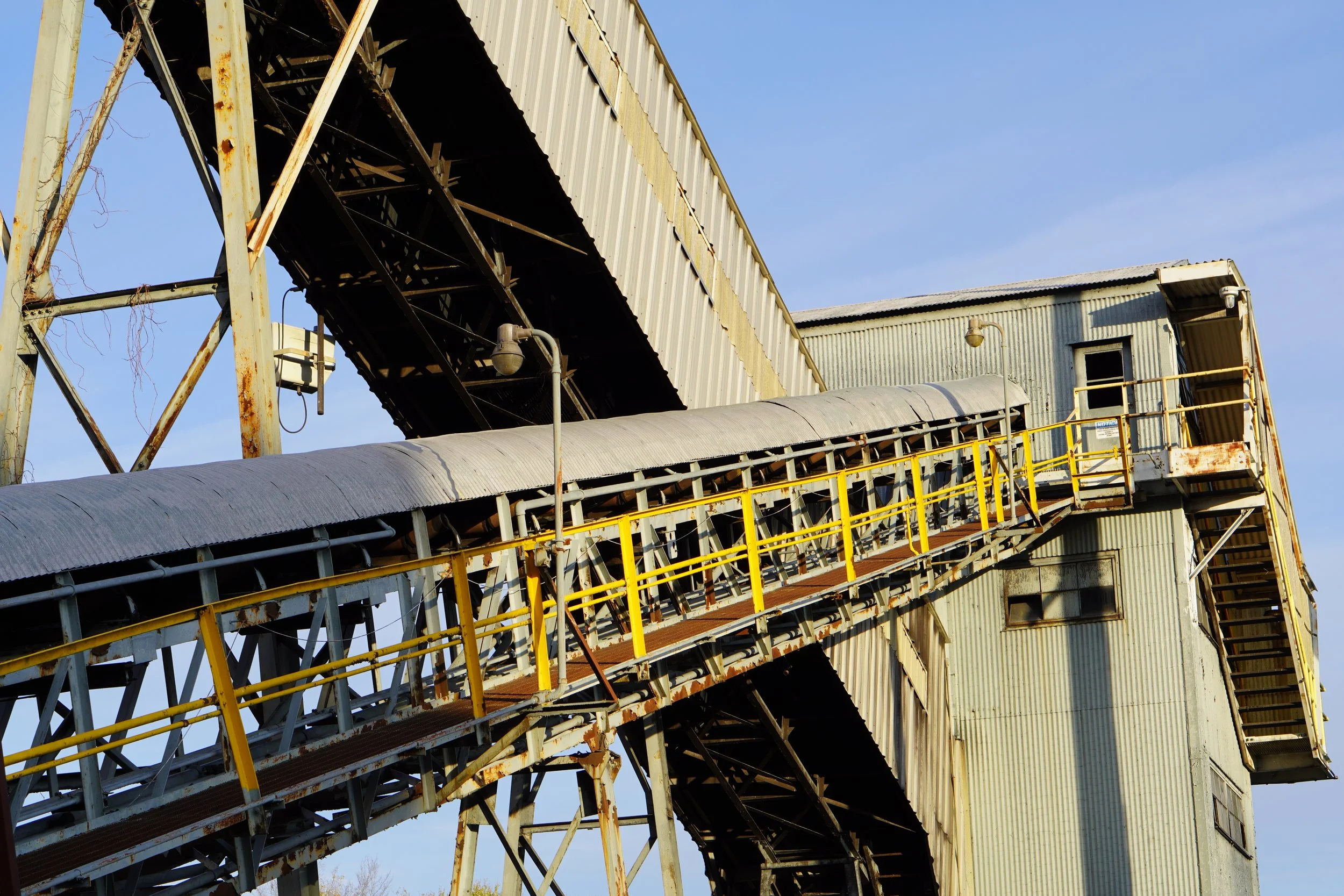 Old industrial building with rusted metal structures, a conveyor system, and yellow safety railings against a blue sky.