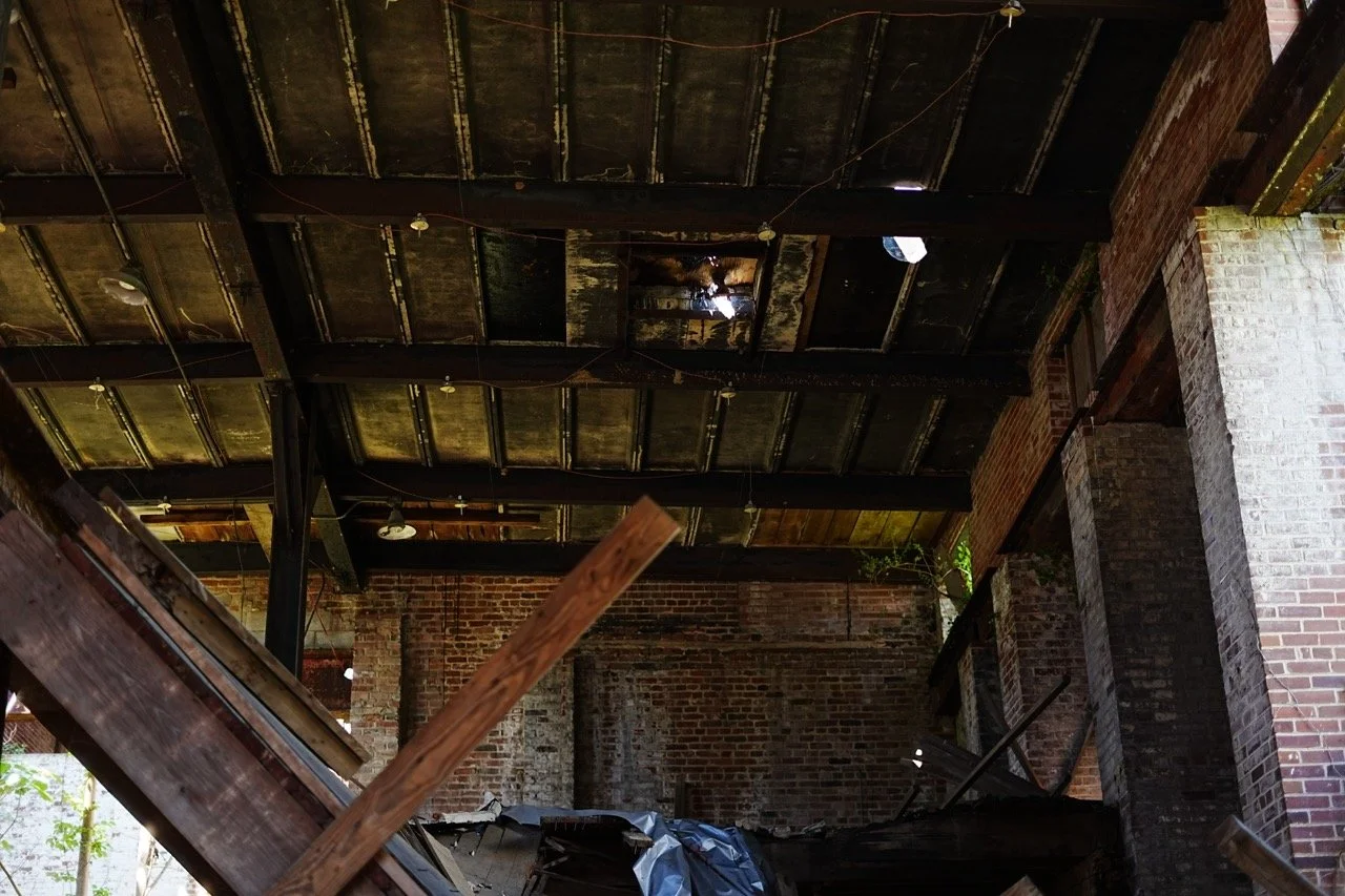 Interior of an abandoned building with exposed brick walls, a deteriorated ceiling with missing panels, scattered debris, and broken furniture.