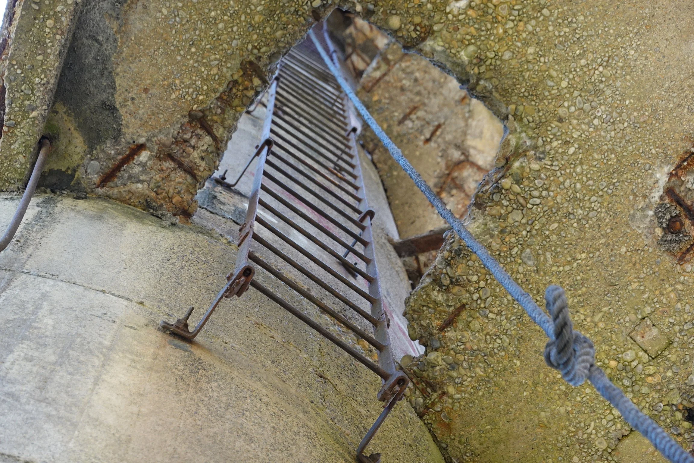 Looking up at the inside of a chimney or tower under construction, showing rough stone walls, metal reinforcements, and a steel ladder attached to the wall with ropes.