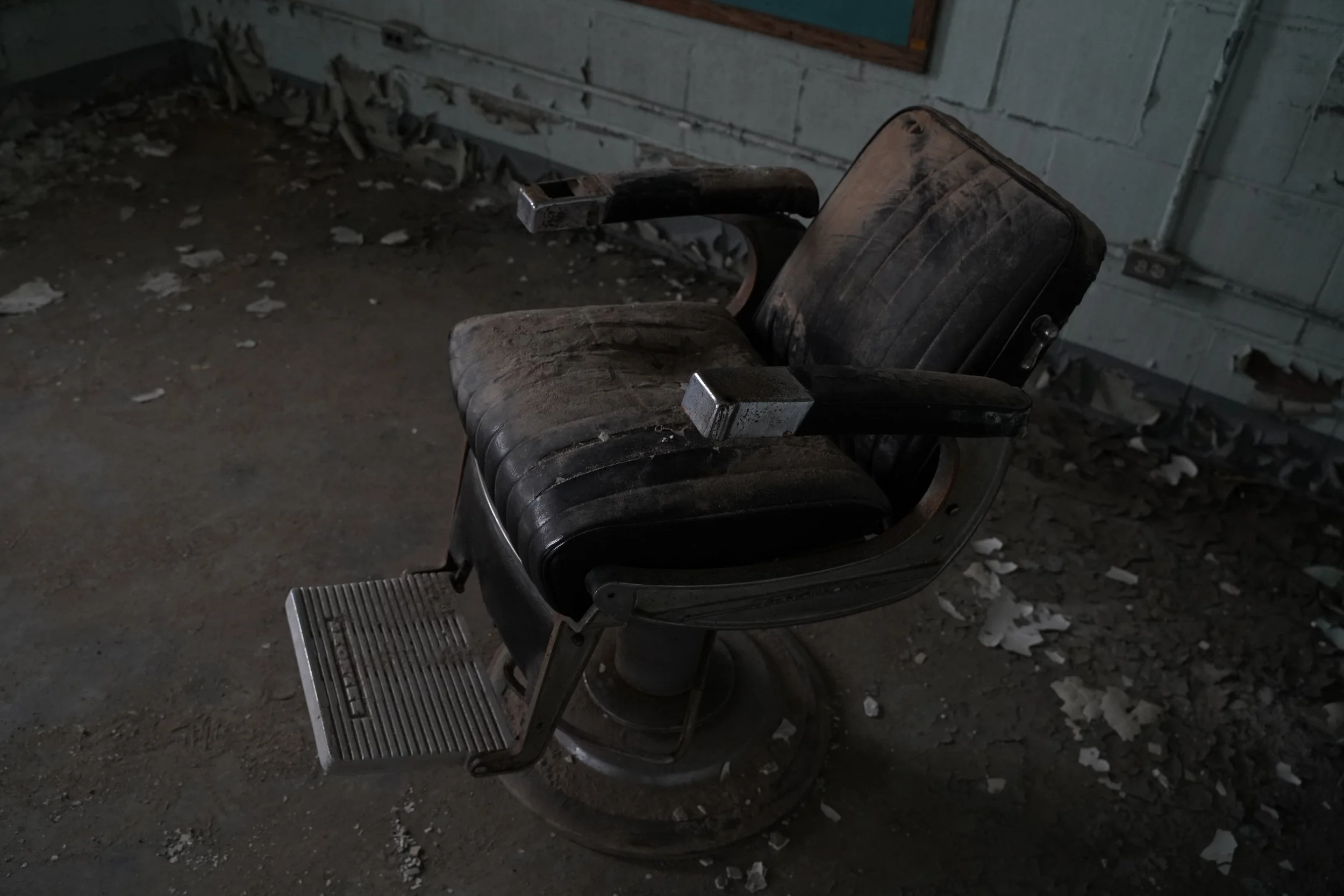 An old, dusty barber chair in a dimly lit, abandoned room with peeling paint and debris on the floor.