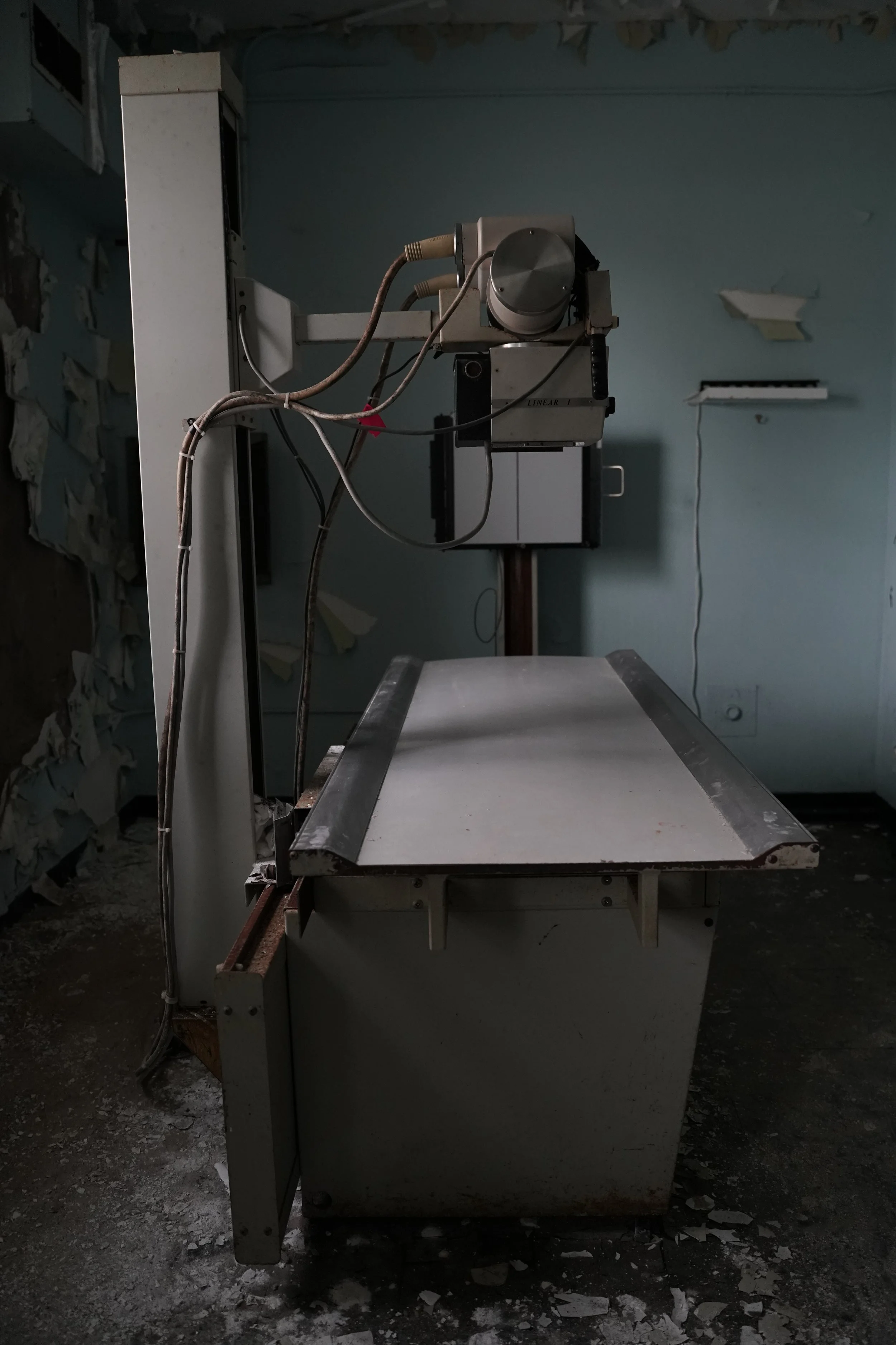 An abandoned X-ray machine in a dimly lit, dusty room with peeling paint on the walls.