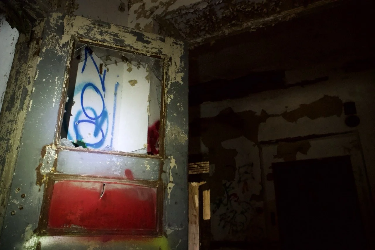 An old, dilapidated doorway with peeling paint and graffiti, leading into a dark, abandoned room with graffiti-covered walls.