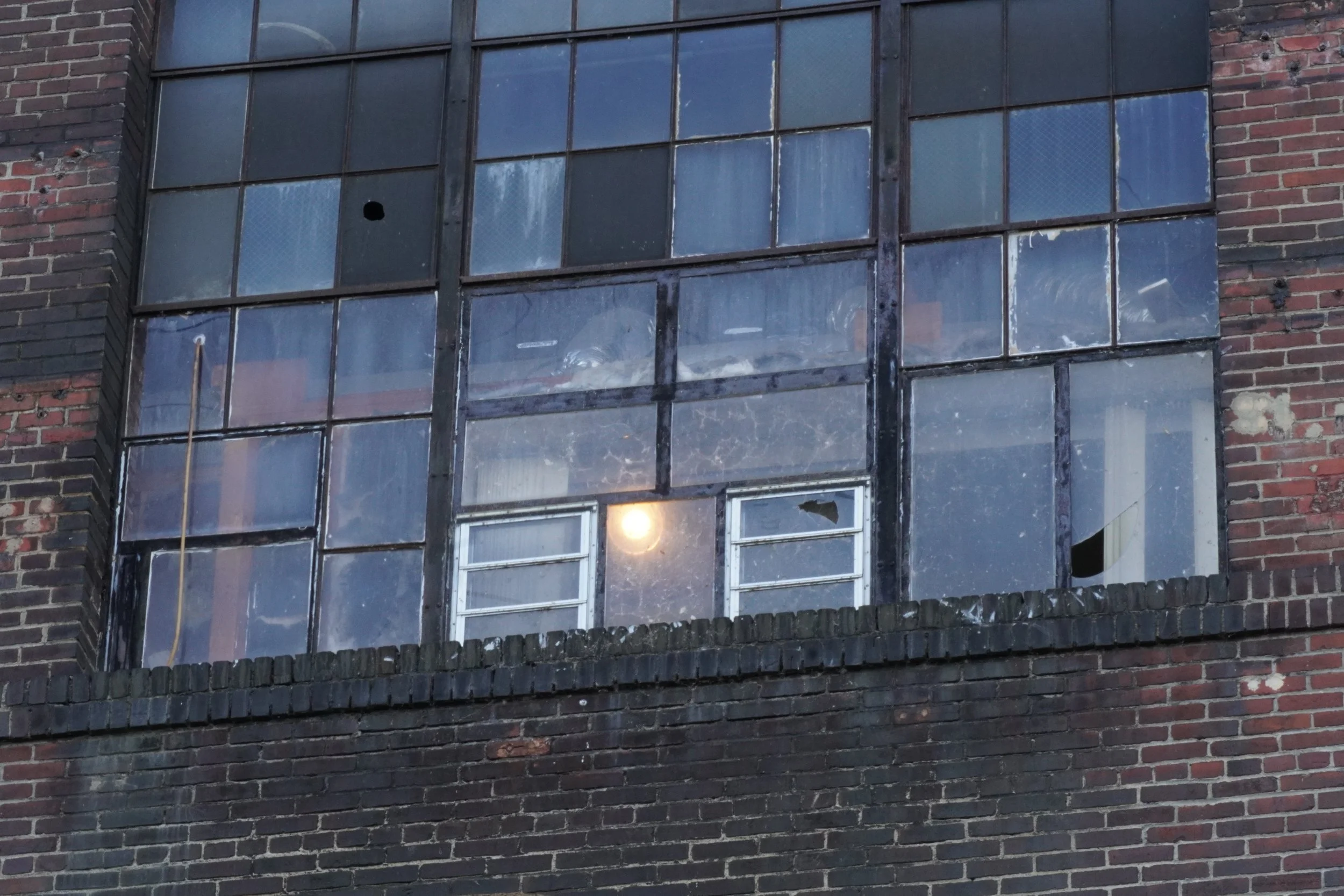 A multi-story brick building with large windows, some of which are broken or missing glass panes, and a black brick ledge at the bottom.