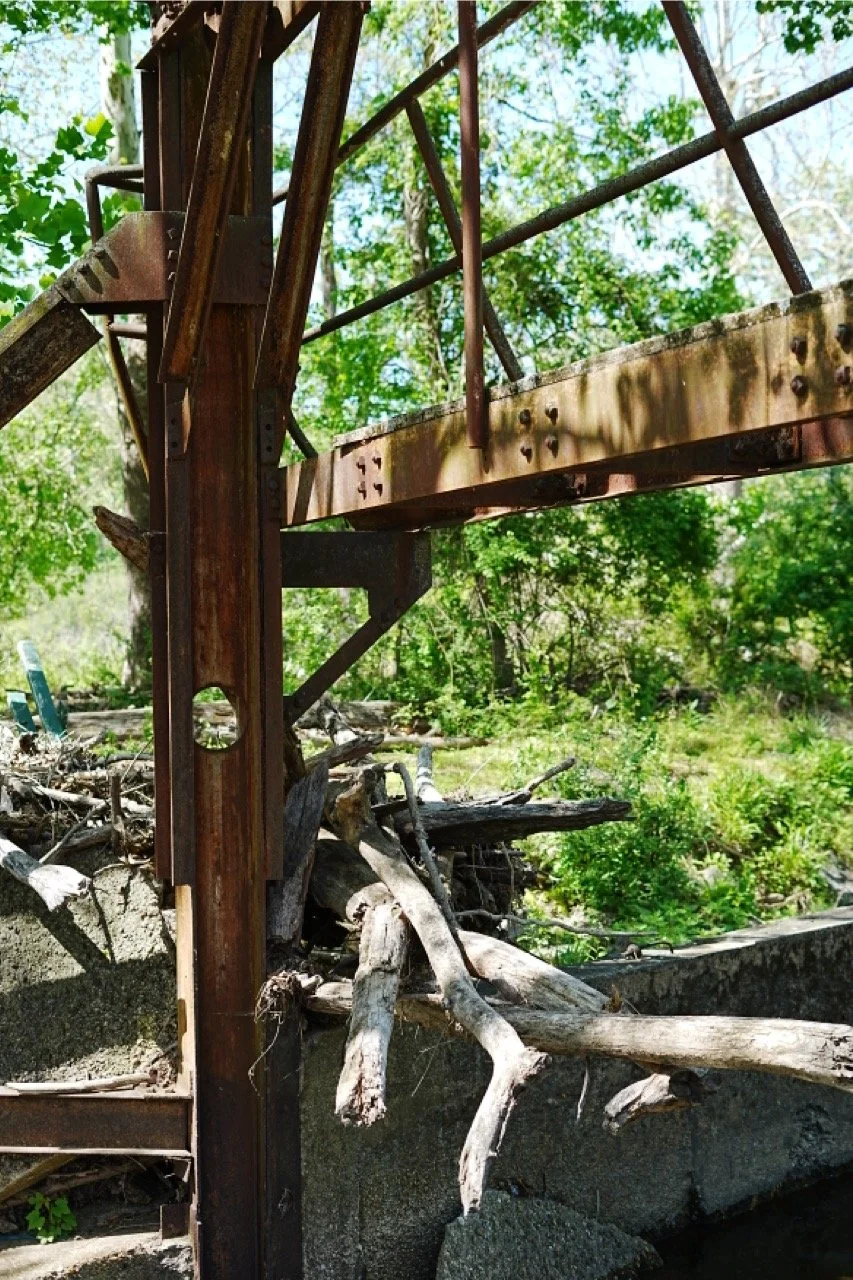 Close-up of a rusty, broken metal structure with wooden debris and overgrown green vegetation in the background.