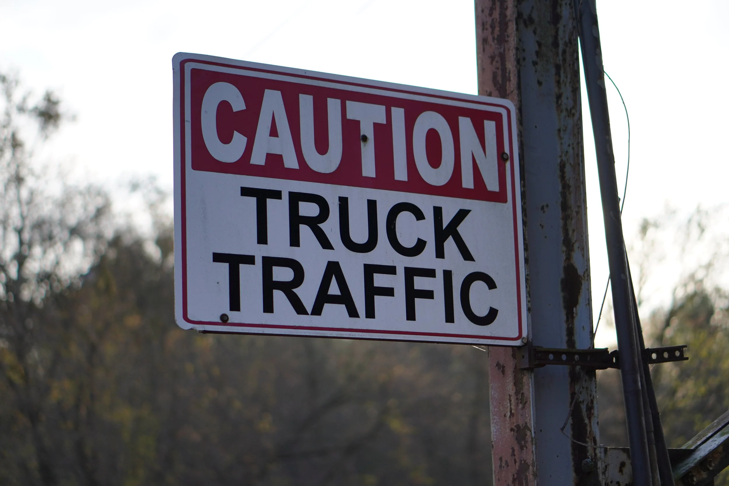 Traffic sign reading 'Caution Truck Traffic' attached to a weathered metal pole with a blurred outdoor background.
