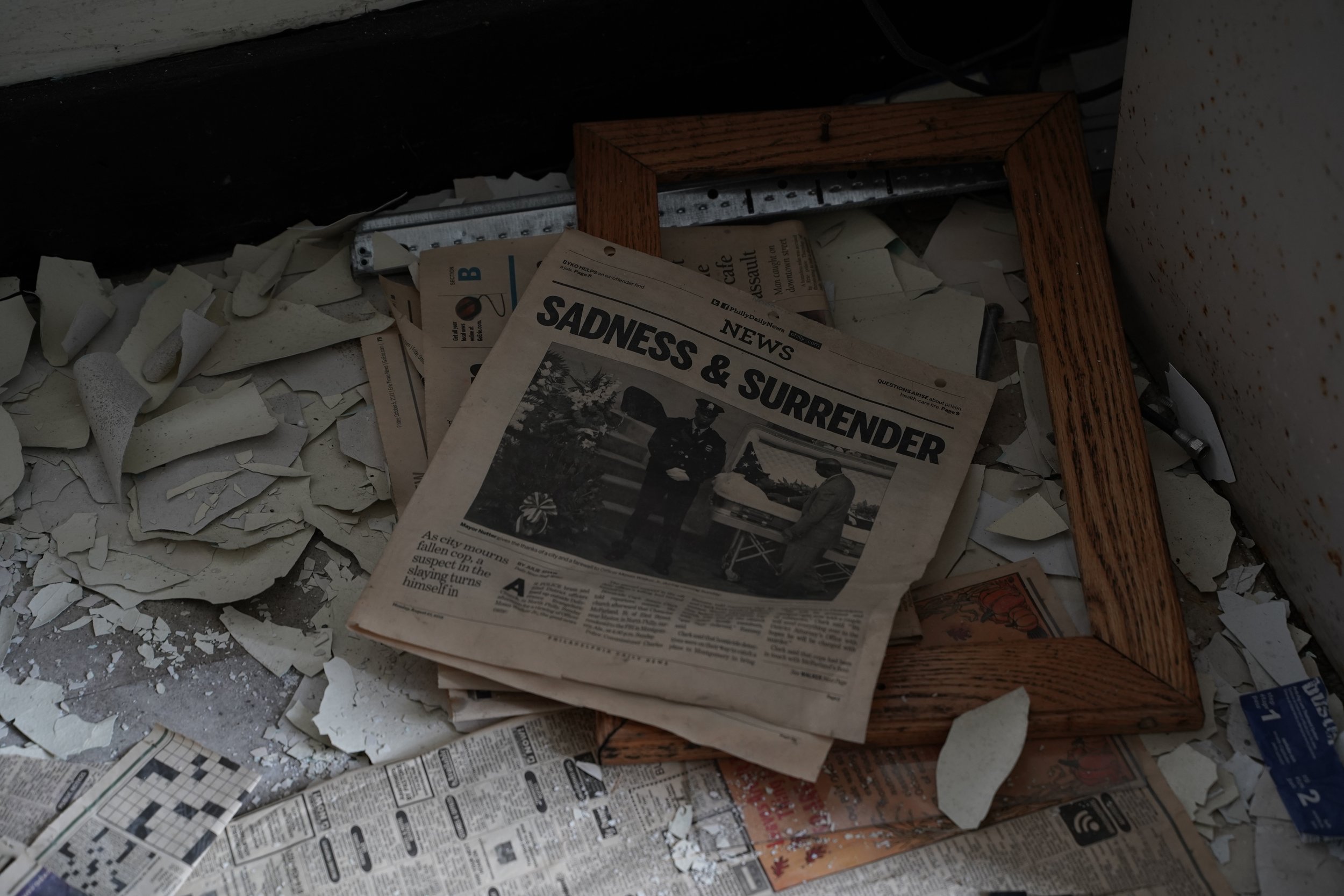 A pile of torn newspapers and trash on the floor with a wooden picture frame leaning against a wall.