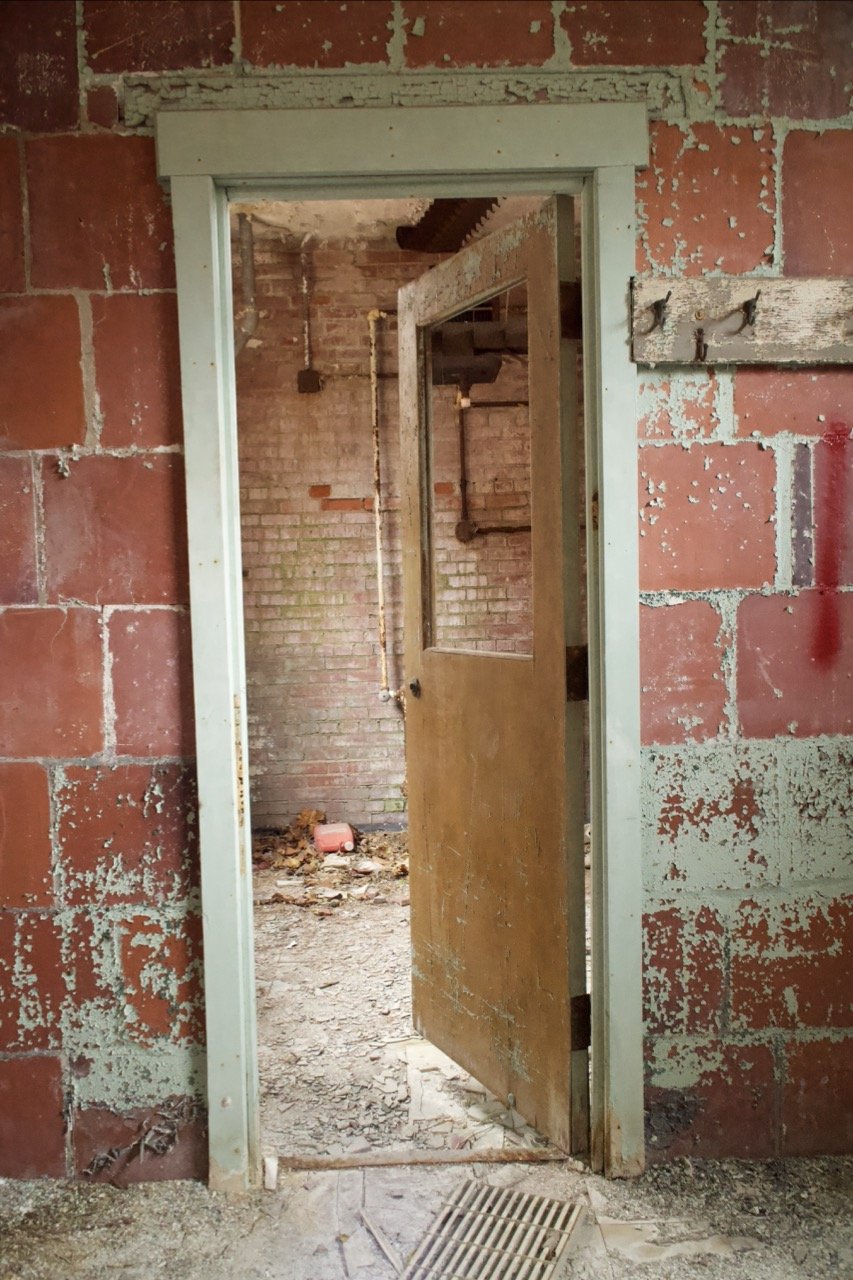 An open doorway reveals an abandoned, dilapidated interior with exposed brick walls, debris on the floor, and a partially visible wooden door inside.