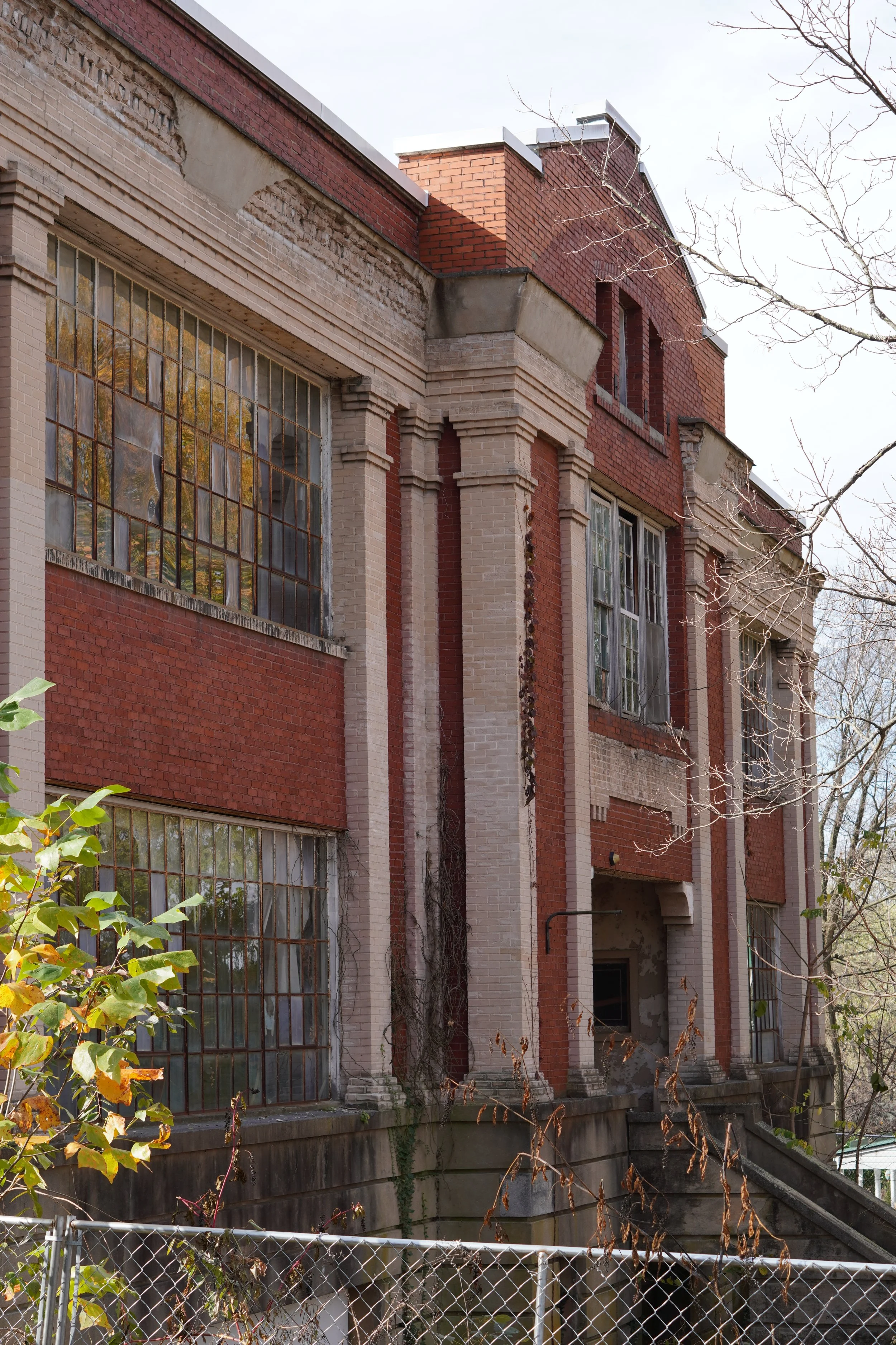 An old, abandoned brick building with large, broken windows and a chain-link fence in front. The building's exterior shows signs of wear and neglect, with some bricks missing or damaged. There are leafless trees nearby, suggesting it is late fall or 