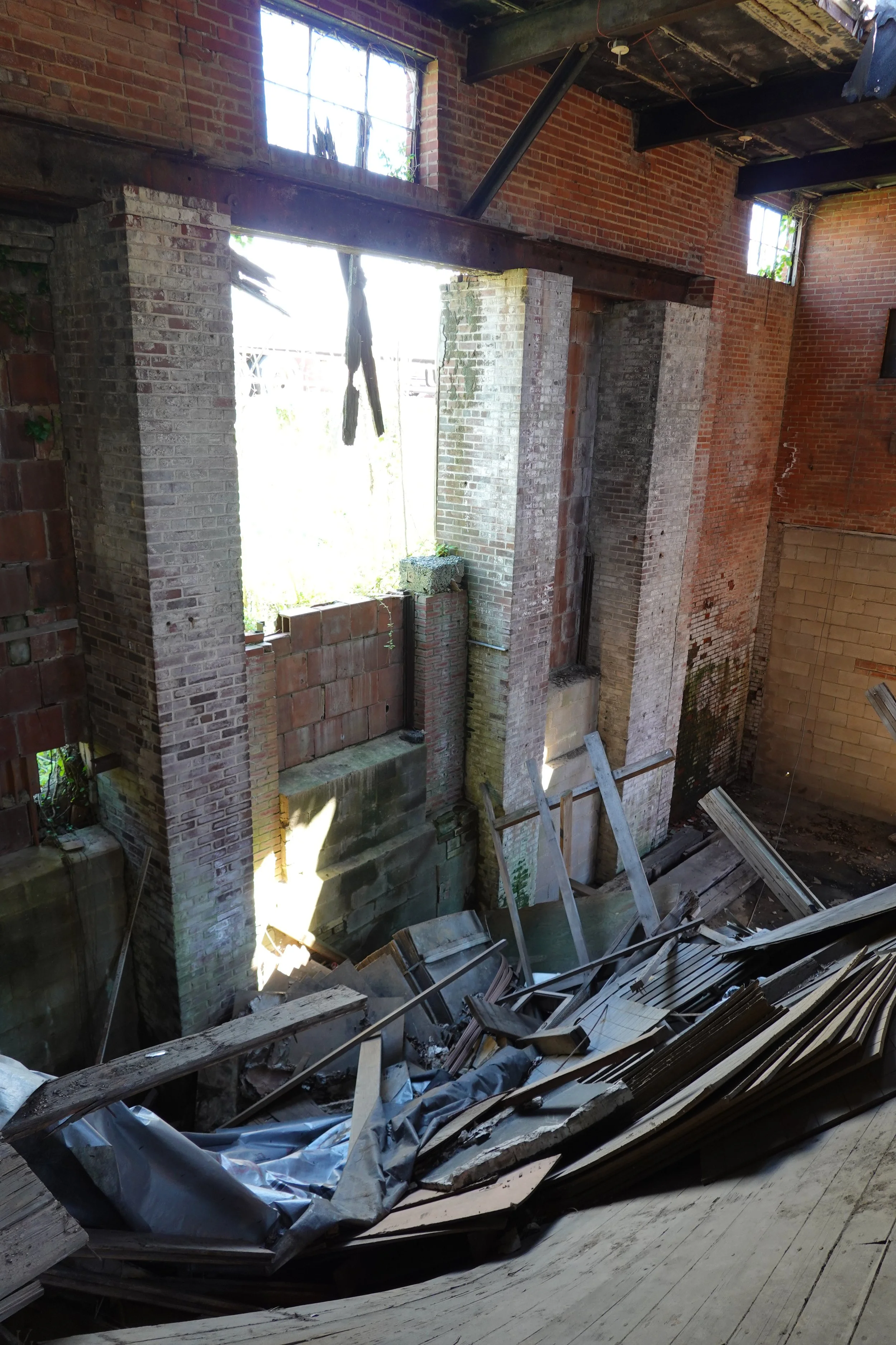An interior space under renovation, with exposed brick walls, large open window spaces, and construction debris including wood, metal, and insulation materials scattered on the floor.
