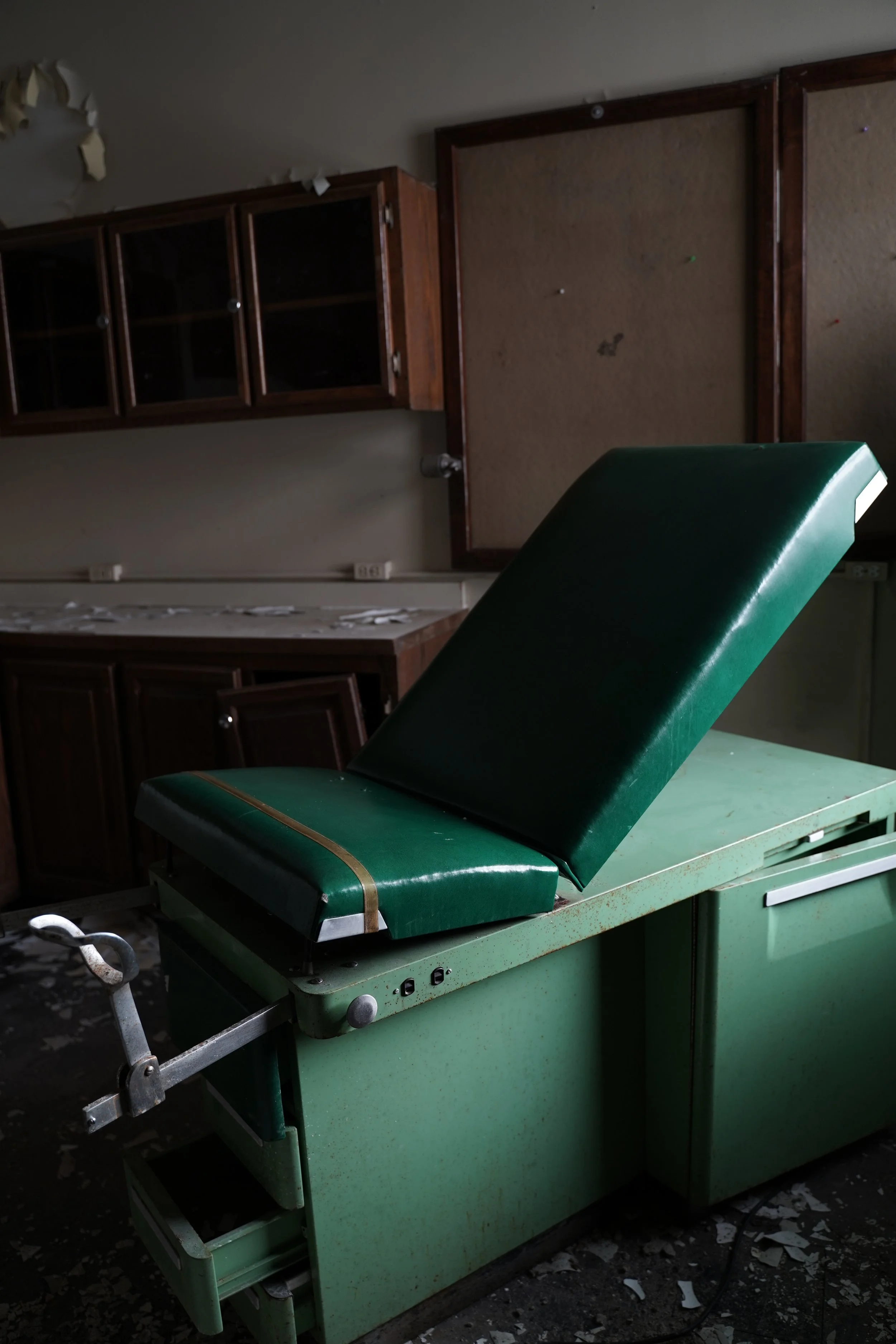 An old, abandoned medical examination table with green padding and a black headrest, set in a dilapidated room with peeling ceiling paint and dirt on the floor.