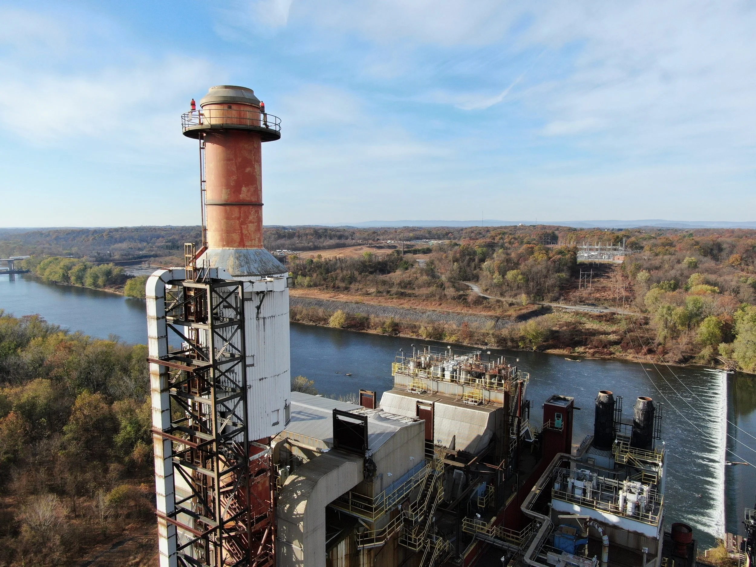 An industrial facility with a large rust-colored smokestack near a river, surrounded by trees and distant hills under a blue sky with scattered clouds.